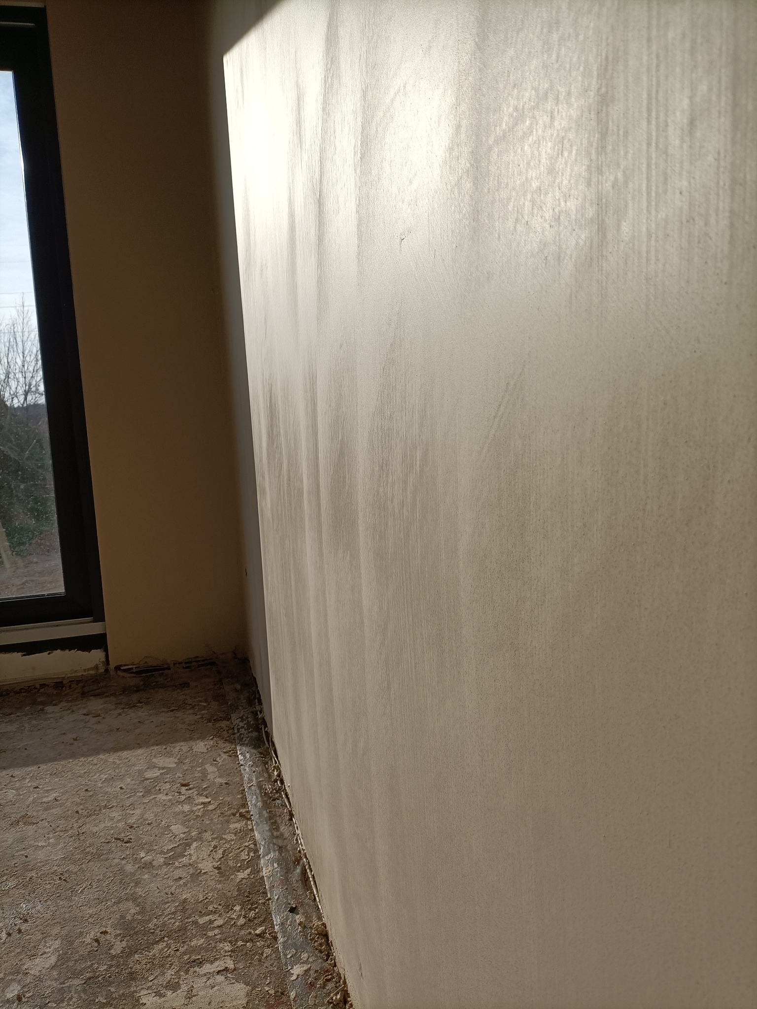 Świeżo nałożona, jasna warstwa gładzi na ścianie w surowym wnętrzu, widoczne ślady po narzędziach i nierówności na powierzchni, okno w tle