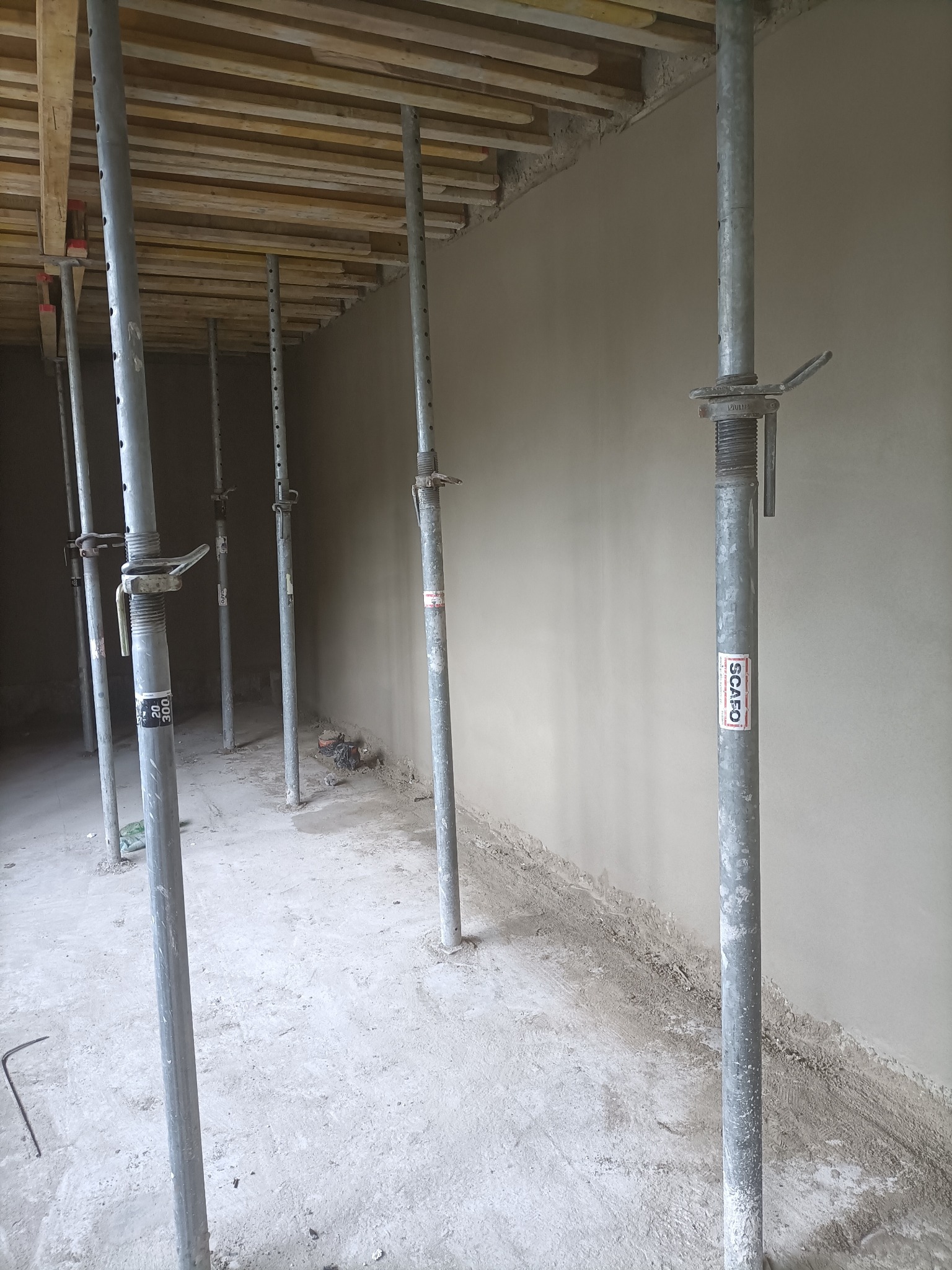 Świeżo otynkowana ściana z widocznymi podporami stropowymi SCAFO, drewnianym szalunkiem stropu i betonową podłogą w surowym stanie.
