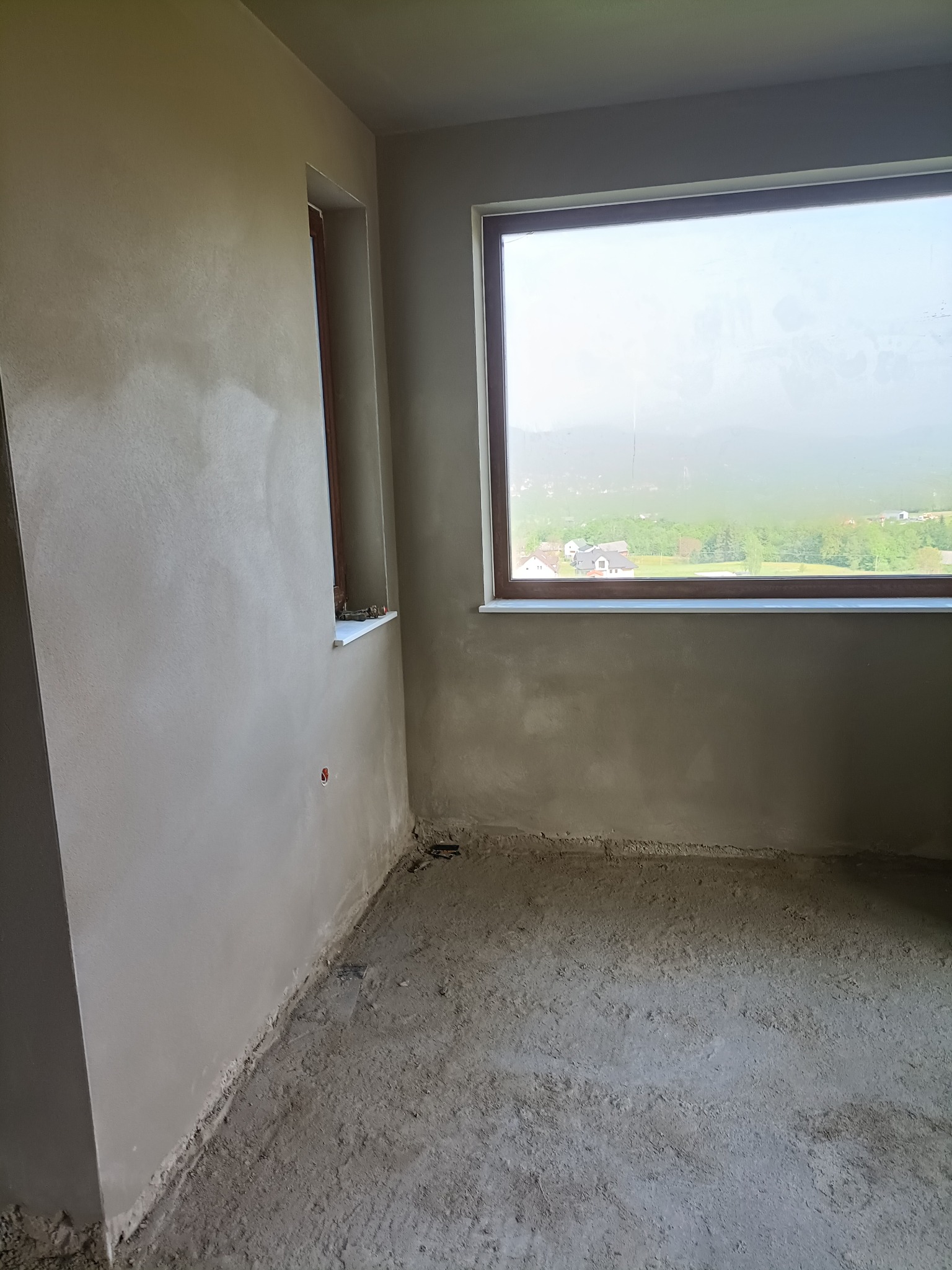 Surowe wnętrze z widokiem na krajobraz przez okno, ściany po tynkowaniu maszynowym, podłoga w trakcie wylewki.