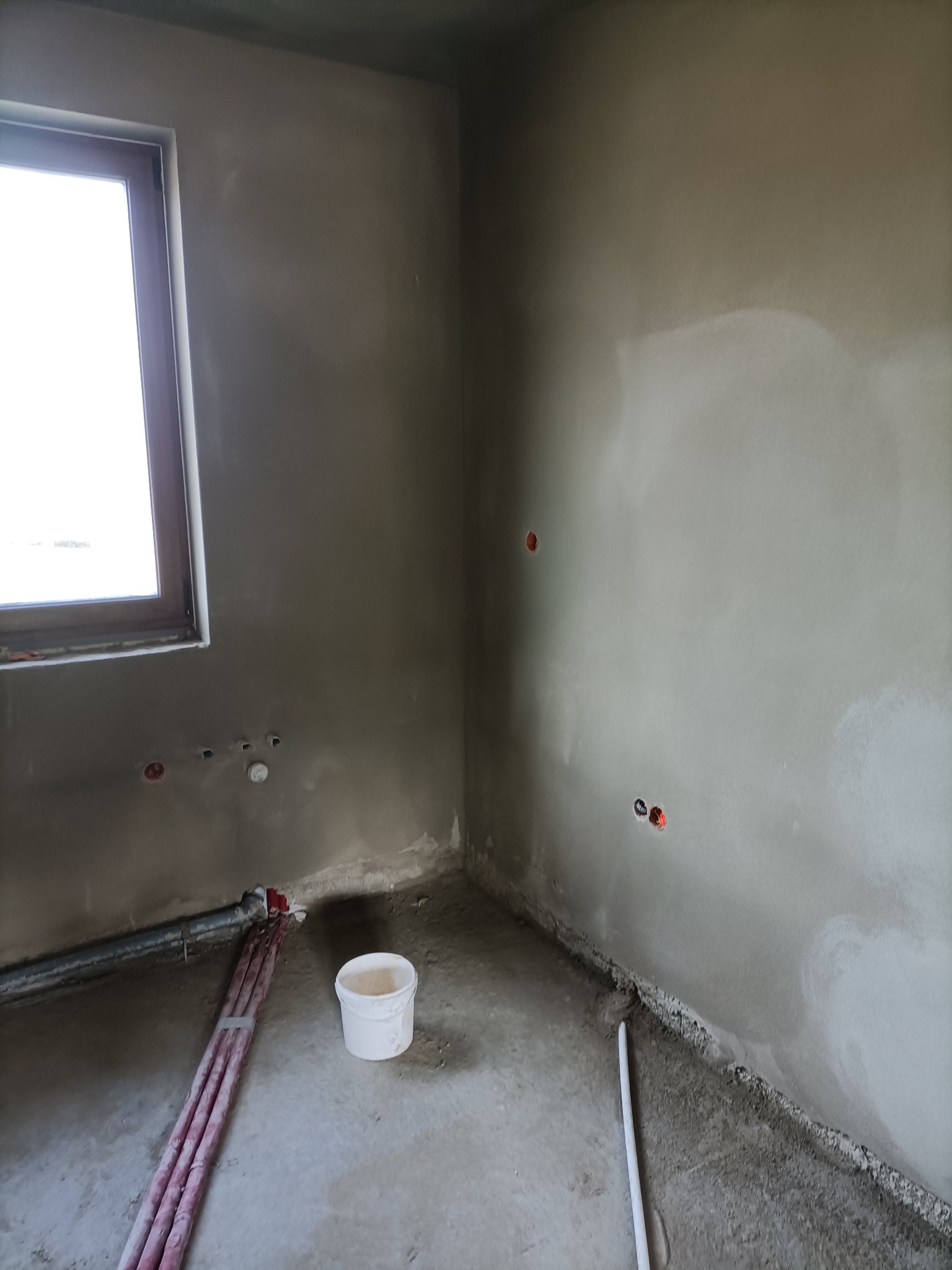 Wnętrze w trakcie prac wykończeniowych: ściany pokryte świeżym tynkiem, widoczne puszki elektryczne, rury grzewcze przy ścianie oraz białe wiadro na podłodze.