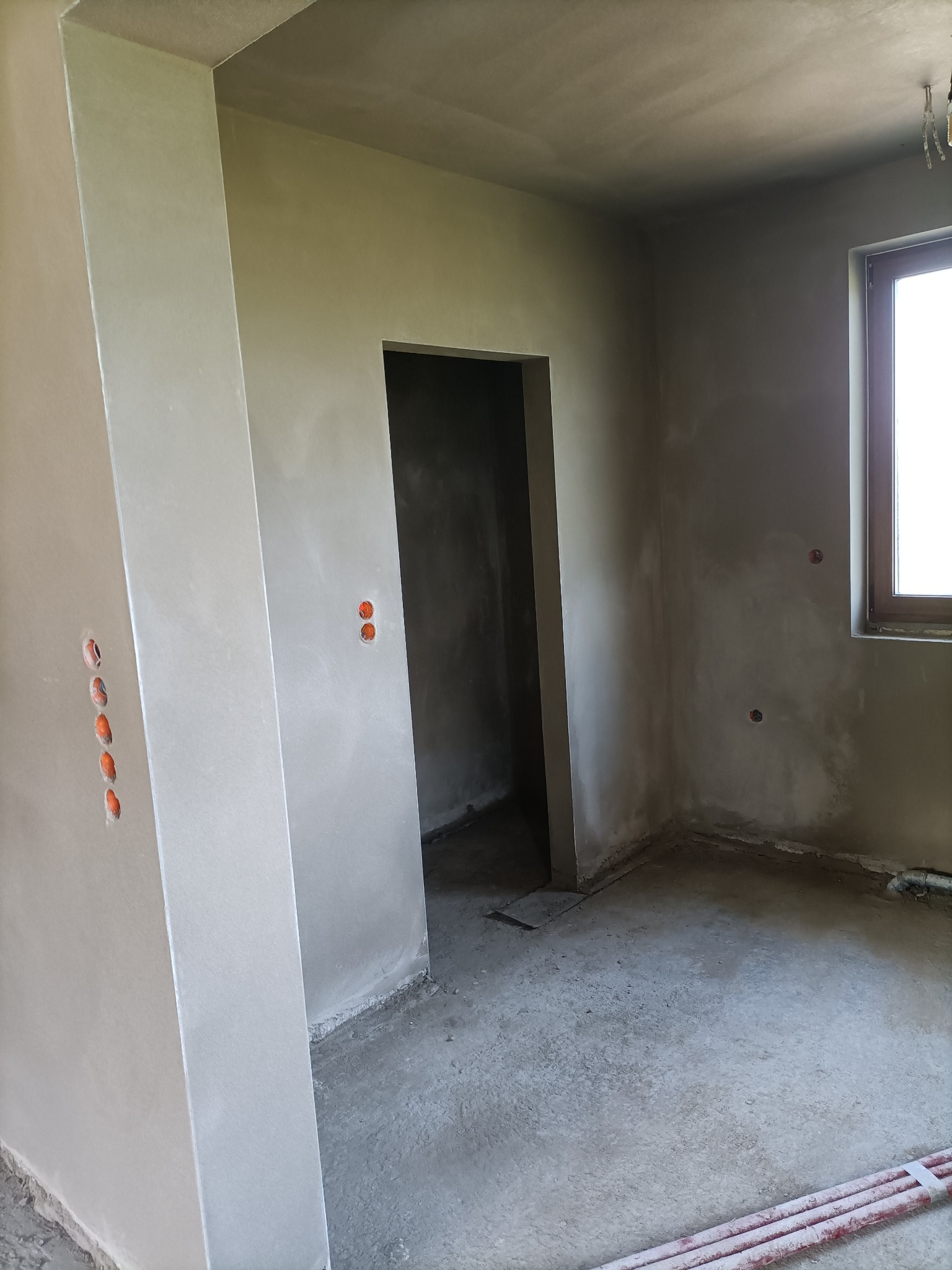Wnętrze w trakcie prac wykończeniowych: świeżo nałożona, jasnoszara warstwa na ścianach i suficie, widoczne puszki elektryczne, surowy beton na podłodze, oraz fragment rur grzewczych w rogu...