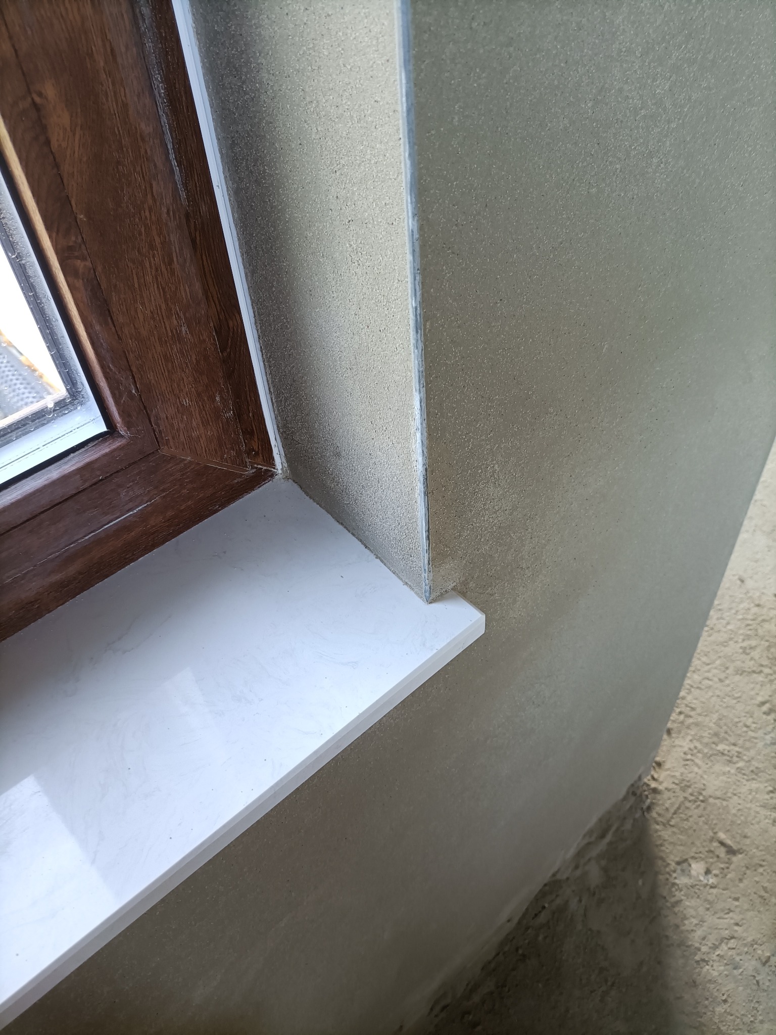 Ściana po nałożeniu tynku maszynowego, widoczny fragment okna z drewnianą ramą i białym parapetem, surowy stan podłoża.