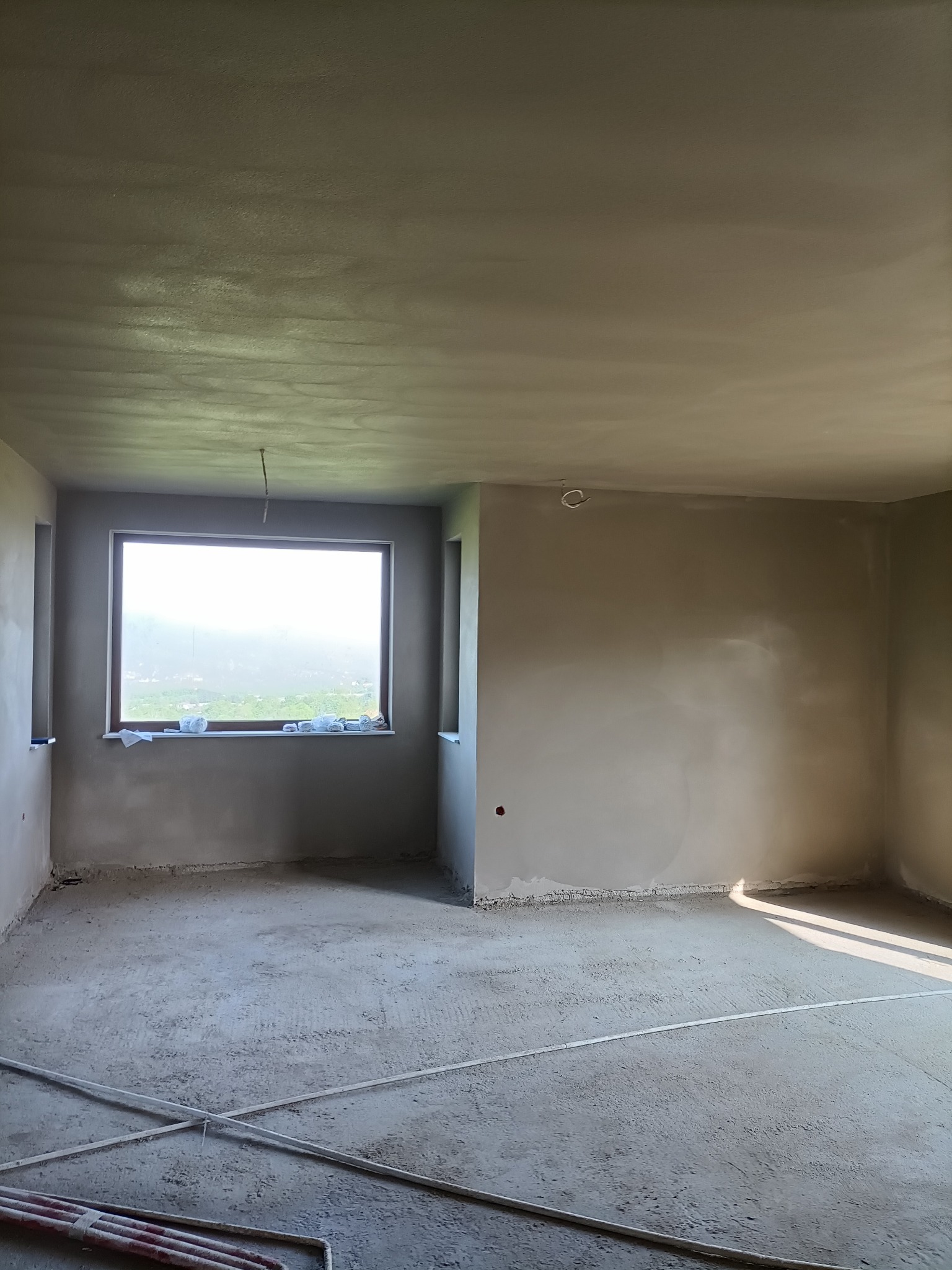 Surowe wnętrze z oknem, ściany i sufit pokryte świeżym tynkiem, na podłodze widoczne rury instalacyjne.