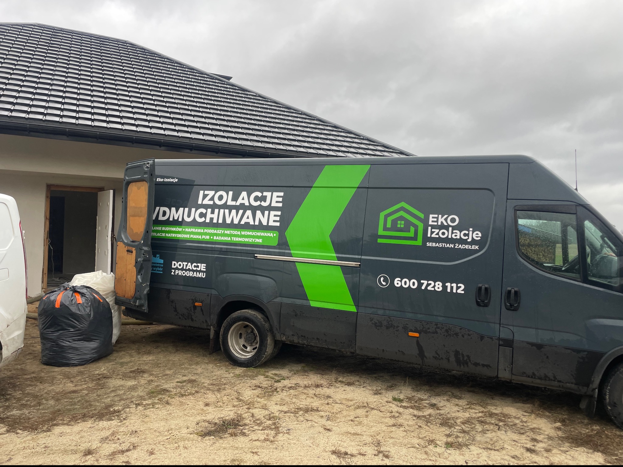 Szary samochód dostawczy z zielonymi elementami graficznymi i napisem 'Izolacje Wdmuchiwane' zaparkowany przed domem w trakcie prac budowlanych, obok worki z materiałem izolacyjnym.