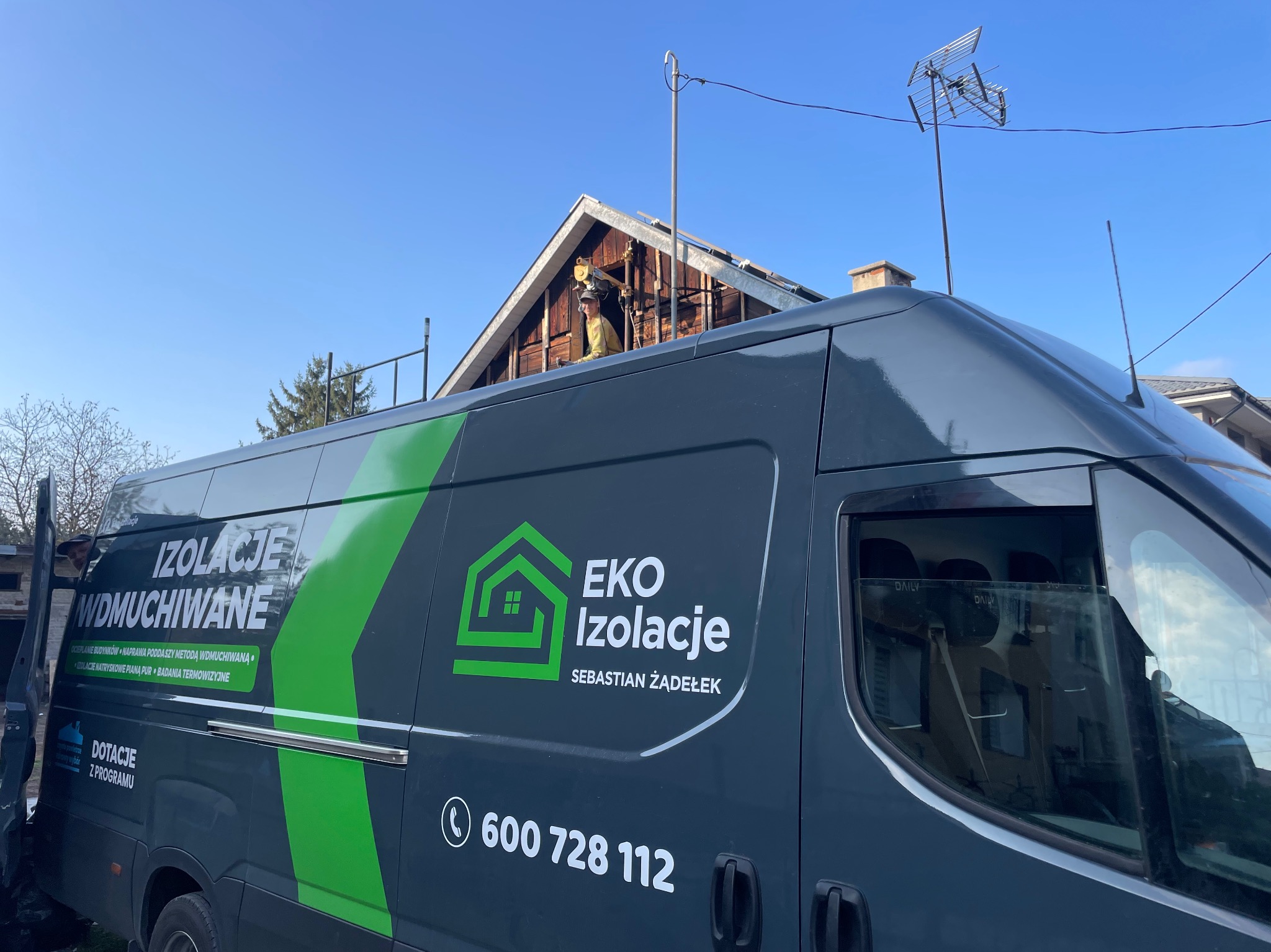 Pracownik w żółtym kombinezonie wykonuje prace na poddaszu drewnianego domu, widoczne elementy konstrukcyjne dachu, na pierwszym planie samochód dostawczy firmy z logo 'EKO Izolacje' i numerem...