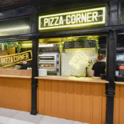Wnętrze stoiska gastronomicznego Pizza Corner z neonowym napisem i logo pizzy, menu na ścianie, piec do pizzy w tle, obsługa za ladą.