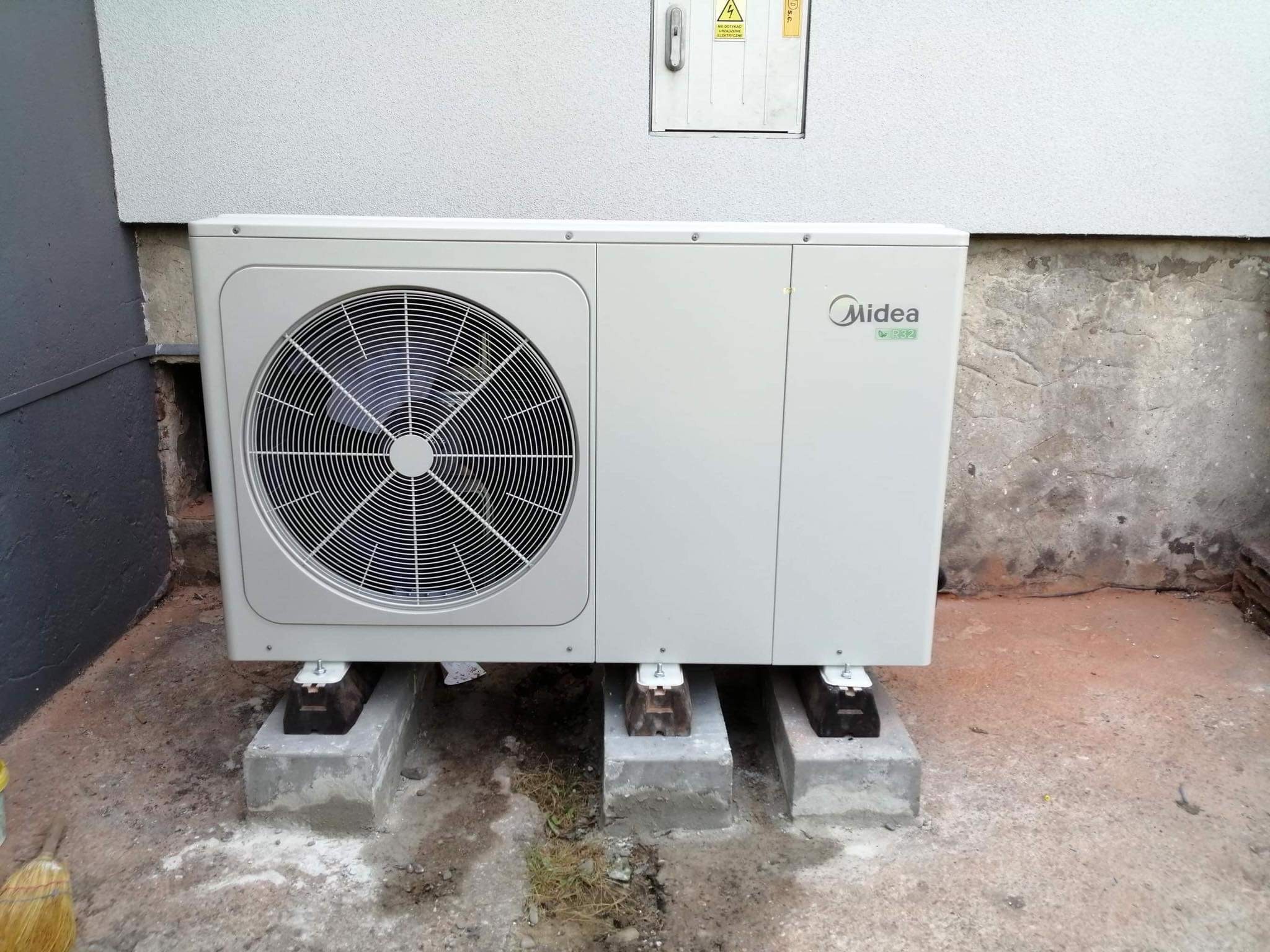 Zewnętrzna jednostka pompy ciepła Midea R32 zainstalowana na betonowych podstawach przy ścianie budynku w Poznaniu, widoczna tabliczka ostrzegawcza na ścianie.