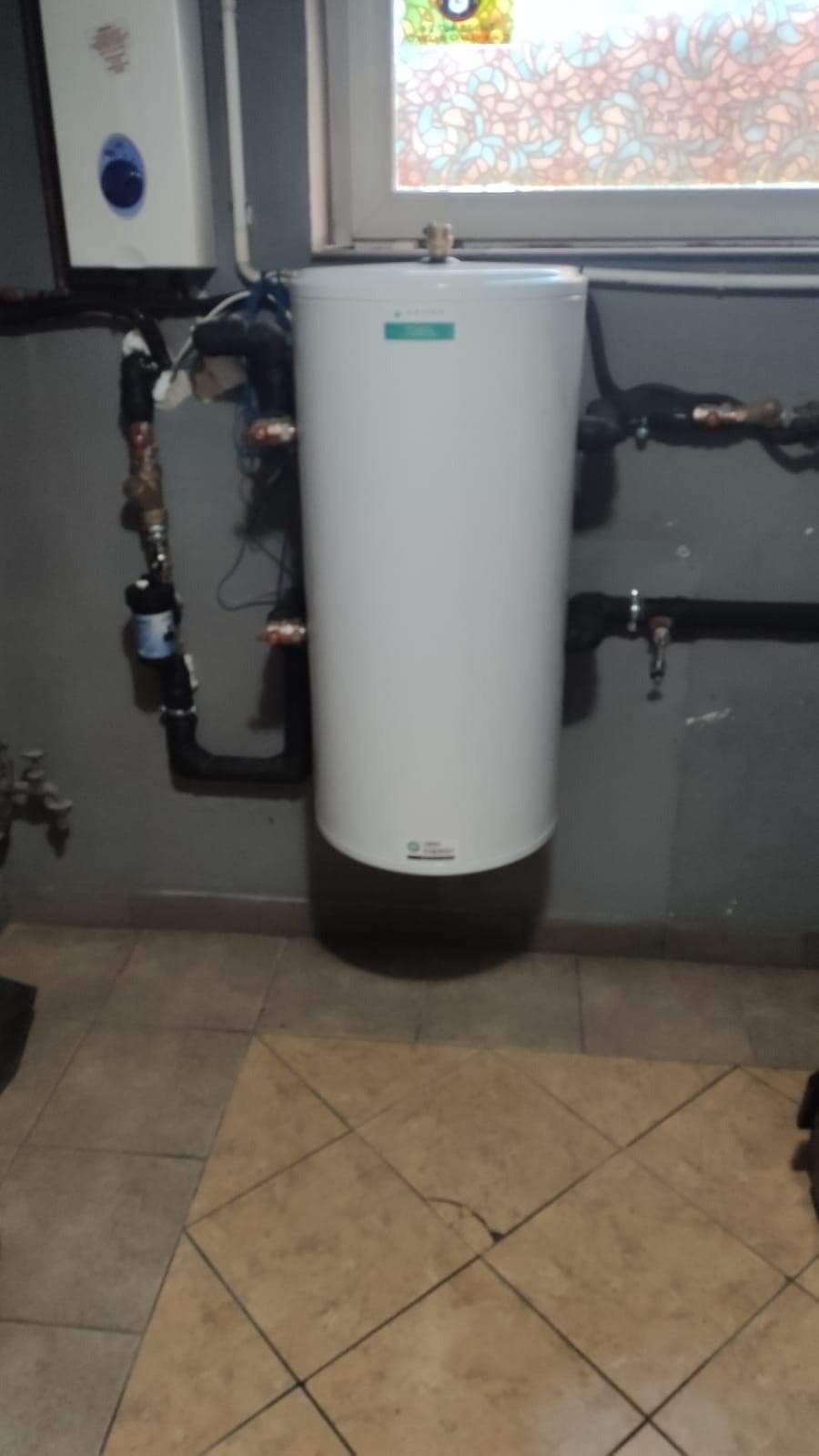 Biały zasobnik ciepłej wody użytkowej z podłączeniami hydraulicznymi, zainstalowany w pomieszczeniu z szarą ścianą i wzorzystym oknem.