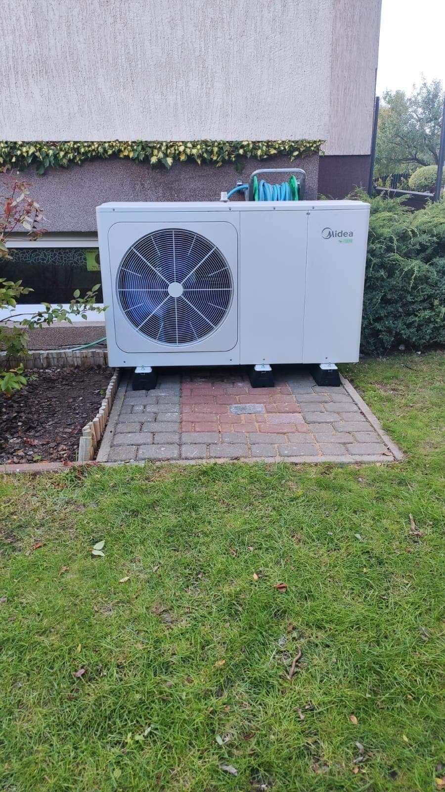 Biała jednostka zewnętrzna z logo Midea zainstalowana na ceglanym podeście w ogrodzie, z zielonym wężem zwiniętym na górze, na tle ściany budynku porośniętej bluszczem.
