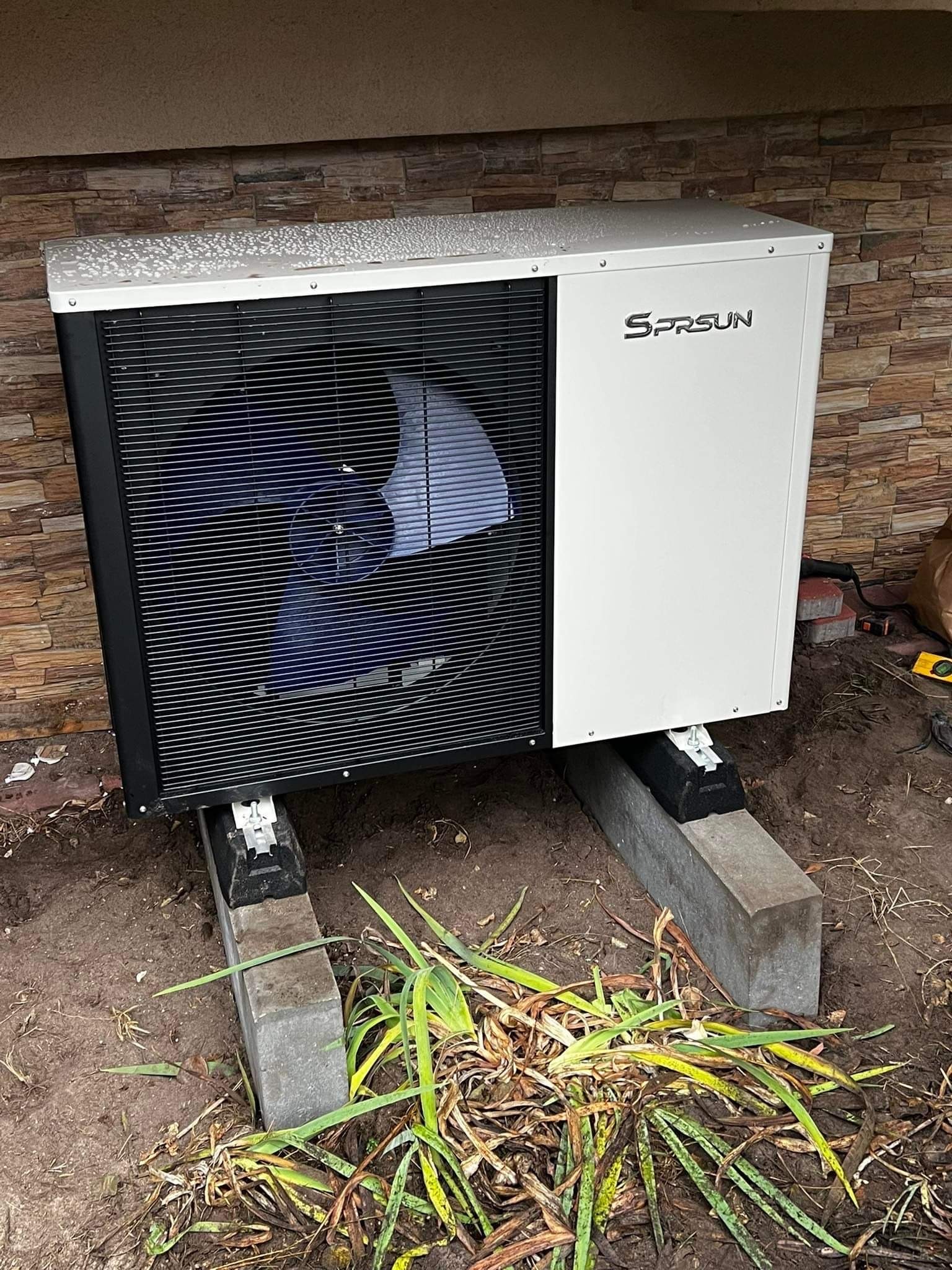 Biała pompa ciepła marki SPRSUN, zamontowana na betonowych podstawach przy ścianie wyłożonej kamieniem dekoracyjnym. Na górnej powierzchni pompy widoczne krople wody, wokół ziemia z resztkami...