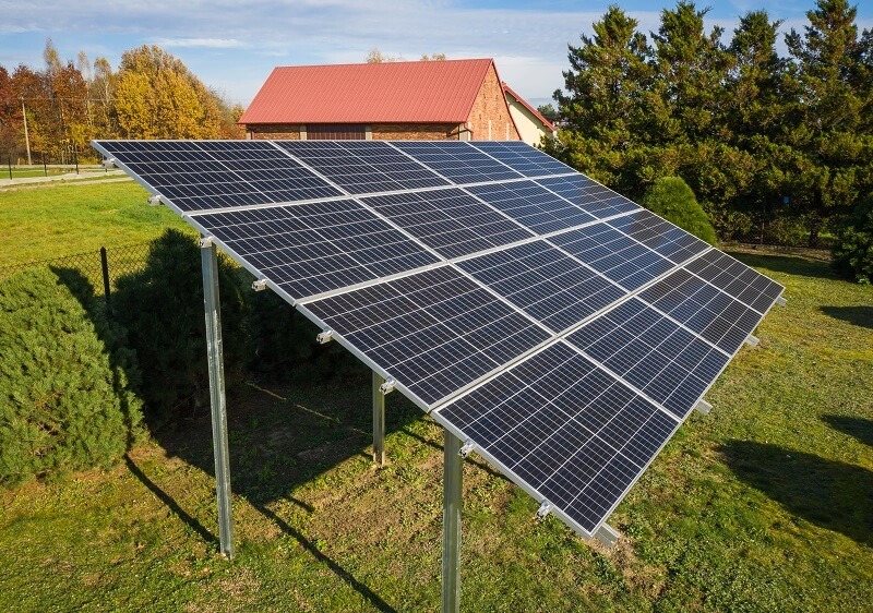 Panel słoneczny na metalowej konstrukcji, ustawiony na trawniku przed domem z czerwonym dachem, widoczne cienie rzucane przez panele.