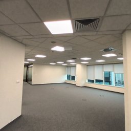 Łukasz Osowiecki Usługi Elektryczne - Oświetlenie biurowe: rastrowe lampy sufitowe w nowoczesnym biurze z szarą wykładziną i oknami z roletami. Widoczne także kratki wentylacyjne.