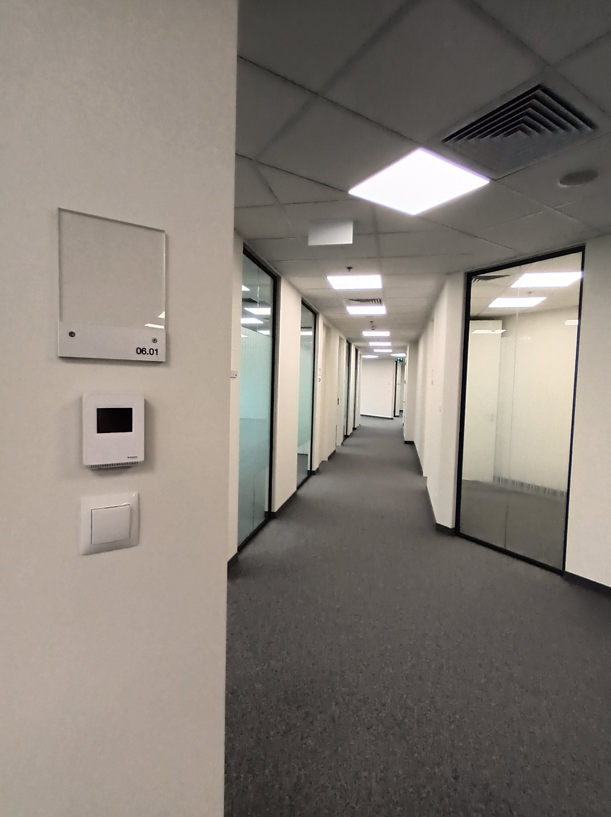 Korytarz biurowy z oświetleniem LED i panelem sterowania. Widoczny włącznik światła. Nowoczesne wykończenie, szara wykładzina, przeszklone biura po bokach.
