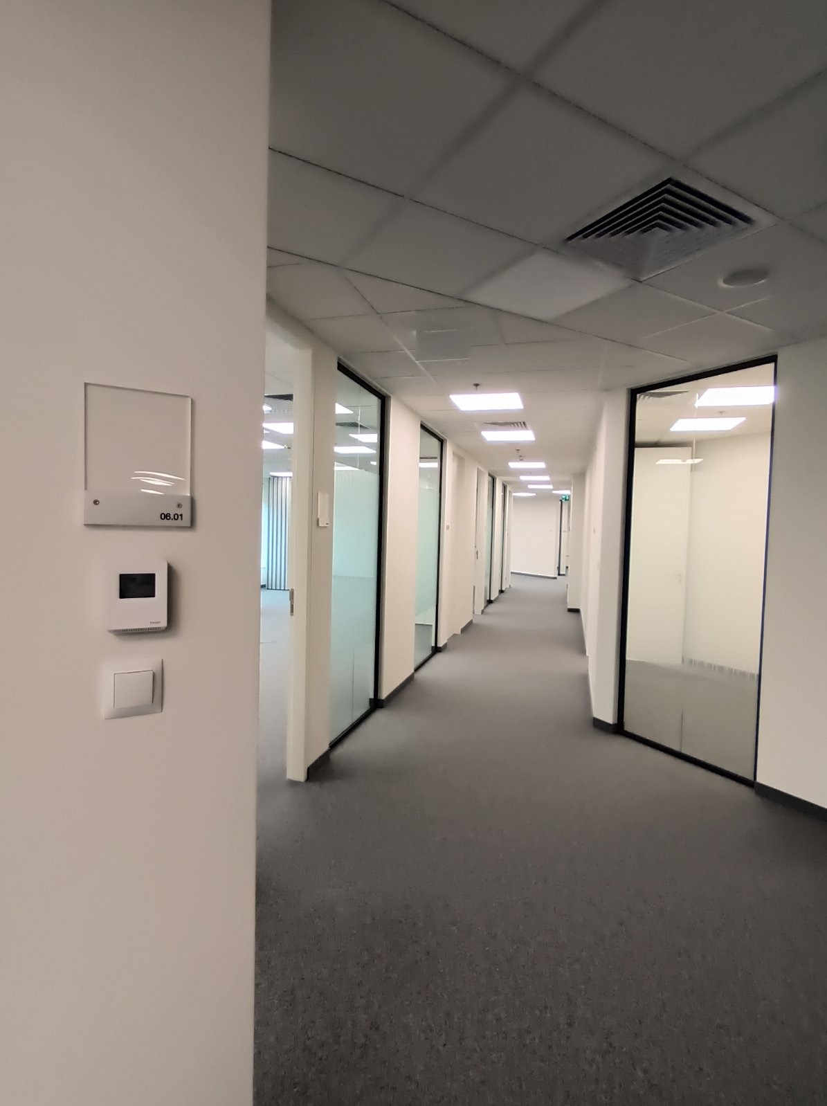 Korytarz biurowy z szarą wykładziną, oświetleniem sufitowym i numerem pokoju 06.01, prezentujący instalacje elektryczne i wykończenie wnętrz w nowoczesnym stylu.