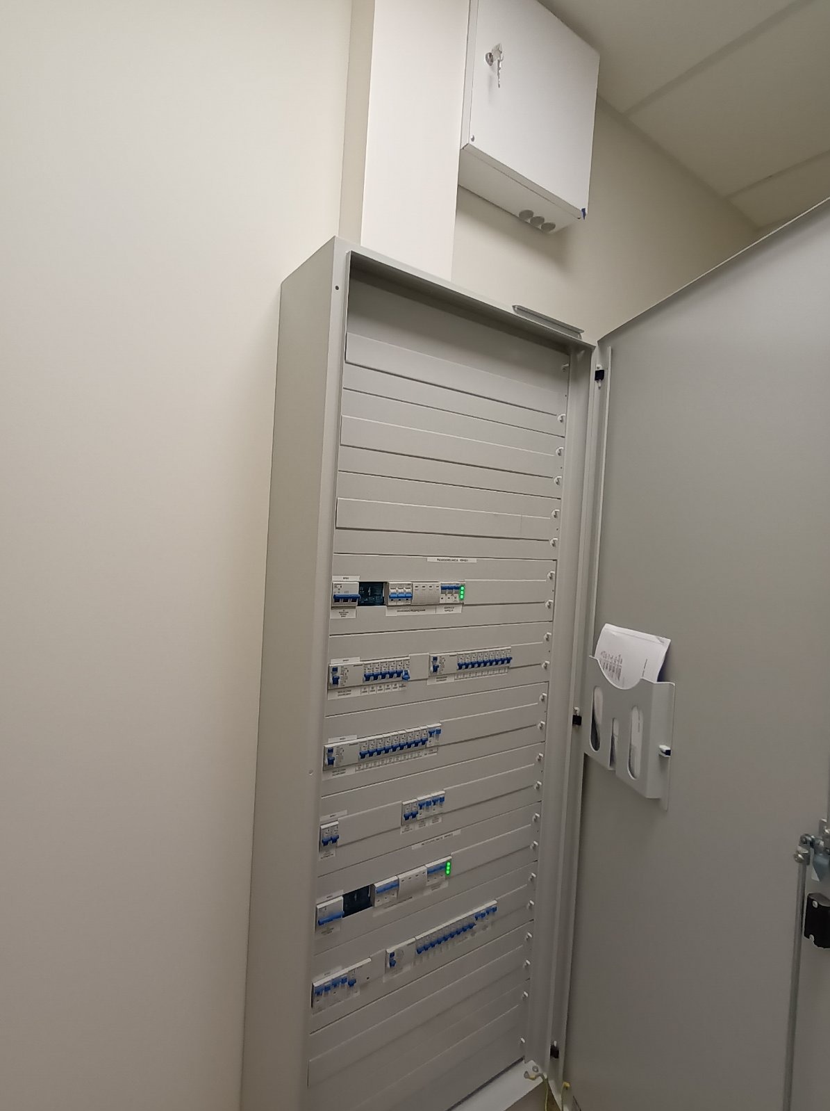 Otwarta, szara szafa rozdzielcza z bezpiecznikami i wyłącznikami, zamontowana na ścianie. Widoczne dokumenty w uchwycie na drzwiach szafy.