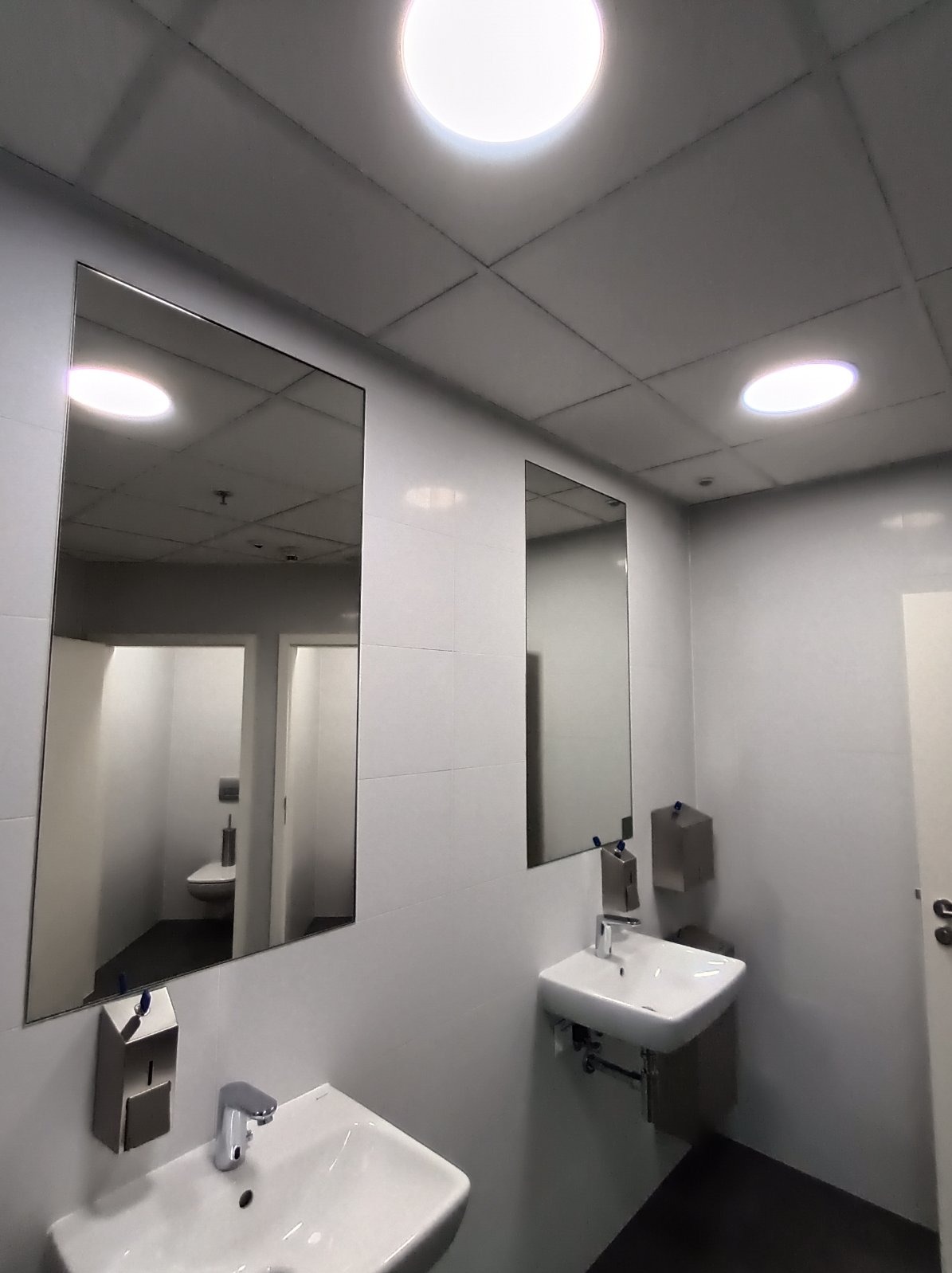 Nowoczesna łazienka publiczna z dwoma umywalkami, lustrami i oświetleniem sufitowym. Widoczne dozowniki mydła i papieru. Minimalistyczny design z białymi płytkami.