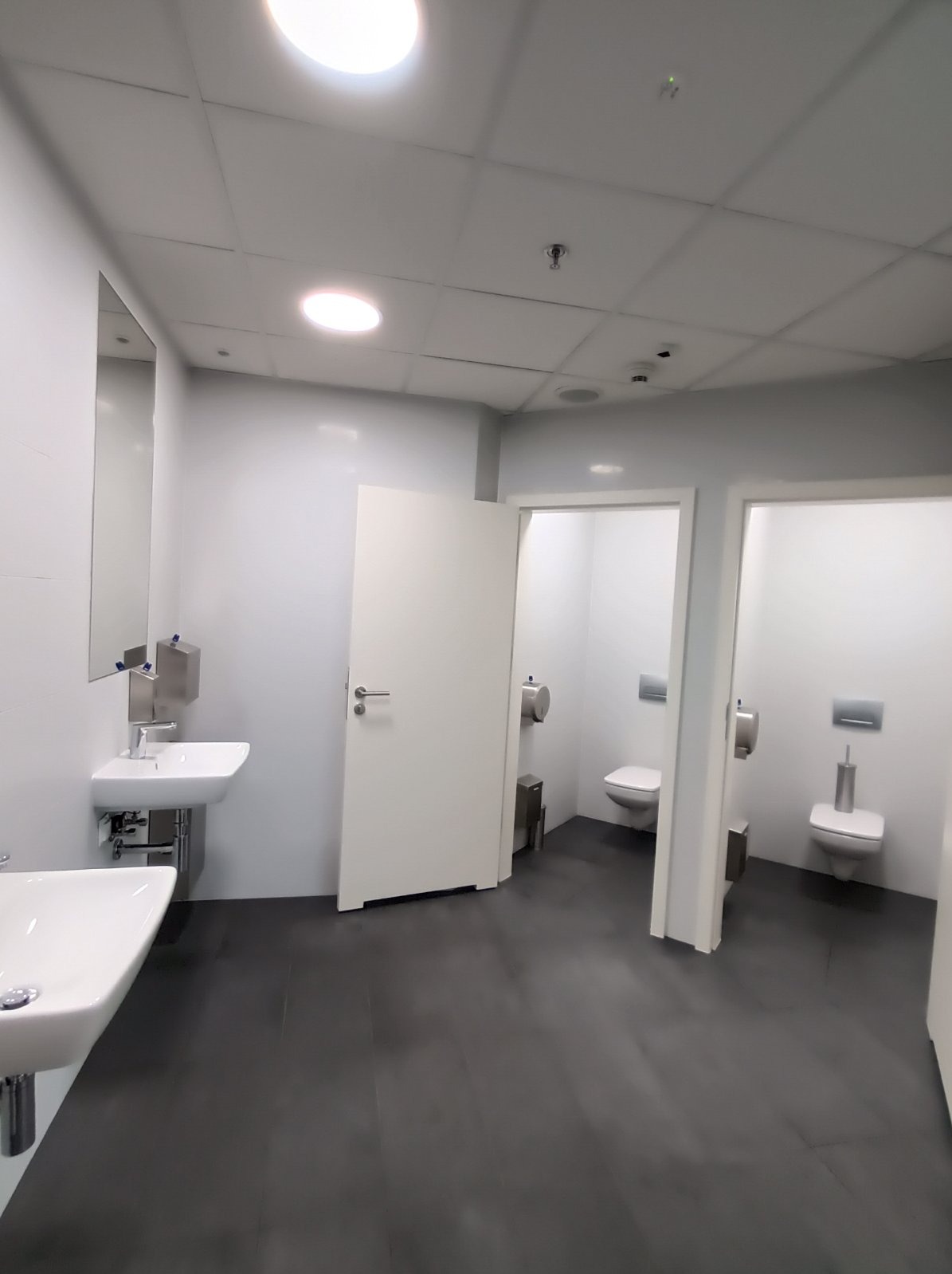 Nowoczesna toaleta publiczna z białymi umywalkami, dozownikami mydła i kabinami WC. Minimalistyczny design, szara podłoga, oświetlenie sufitowe.