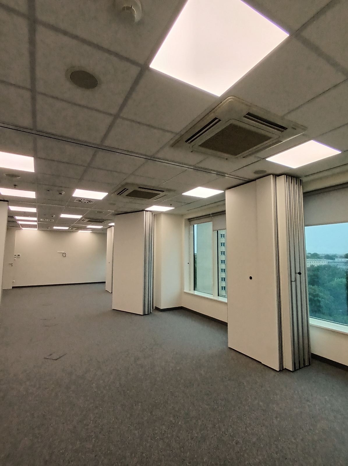 Wnętrze biurowe z podwieszanym sufitem, oświetleniem LED, klimatyzacją i przesuwnymi ściankami działowymi. Widoczne okna z widokiem na zieleń miejską.