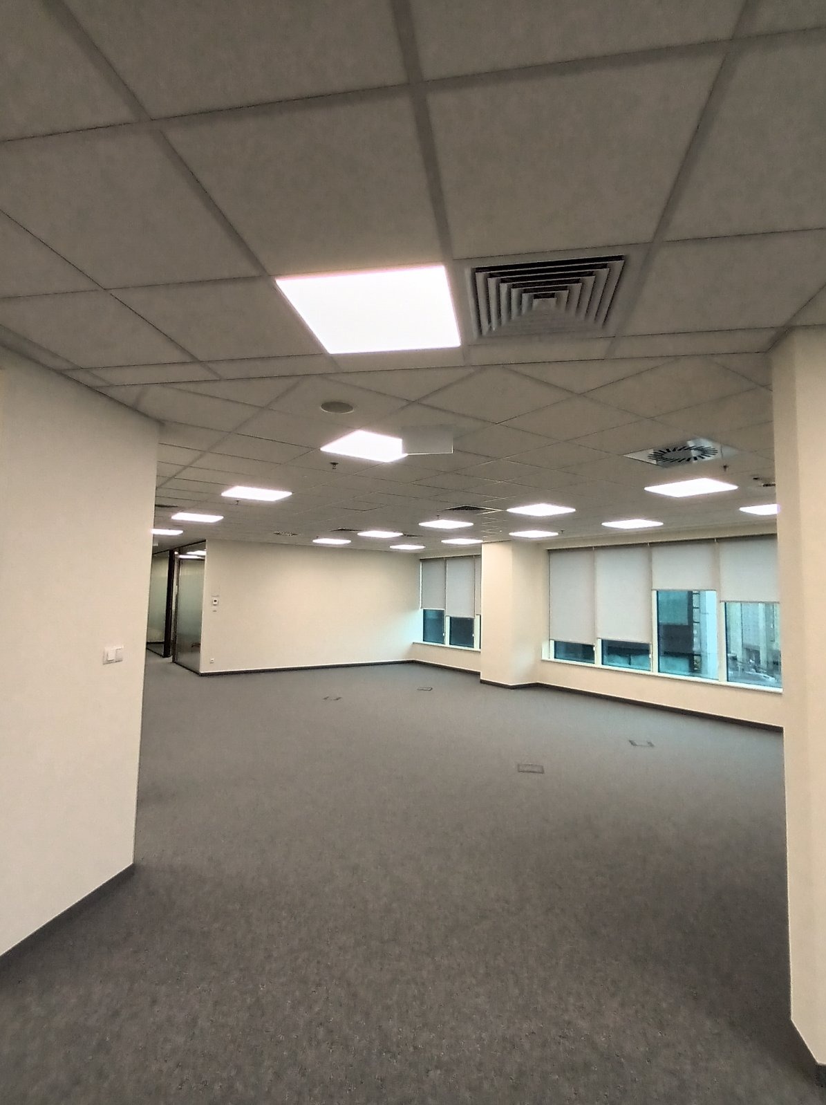 Oświetlenie biurowe: rastrowe lampy sufitowe w nowoczesnym biurze z szarą wykładziną i oknami z roletami. Widoczne także kratki wentylacyjne.