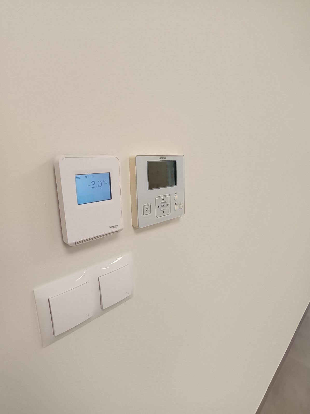 Białe panele sterowania klimatyzacją Hitachi i Schneider oraz włącznik światła na kremowej ścianie. Widoczna niska temperatura na wyświetlaczu.