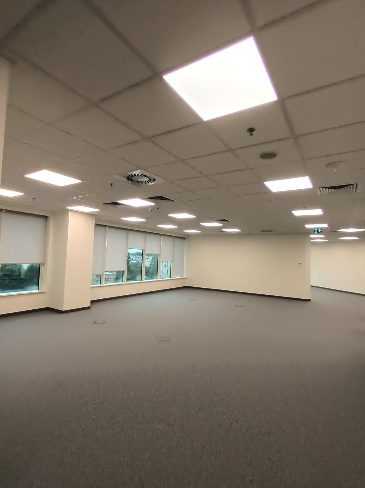 Biuro z oknami i podwieszanym sufitem z panelami LED. Widoczne kratki wentylacyjne i czujniki. Pusta przestrzeń biurowa z szarą wykładziną.