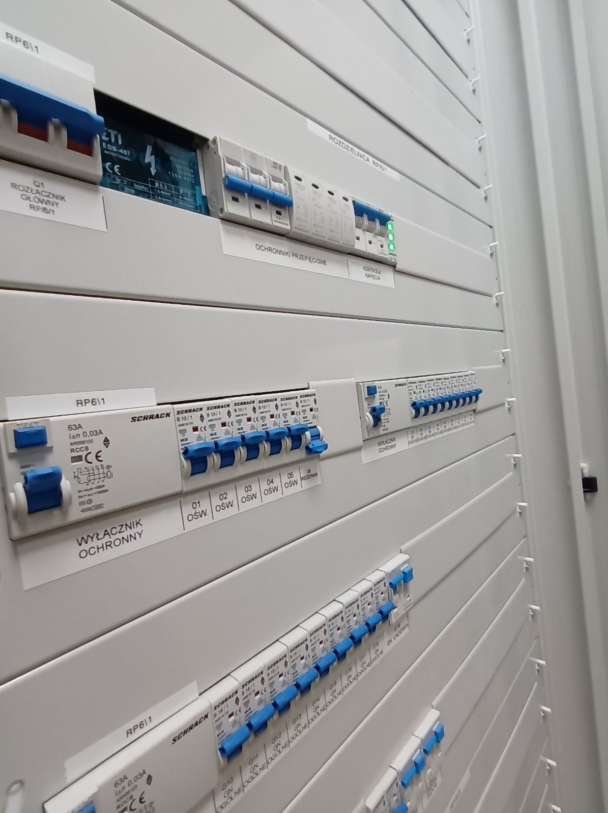 Rozdzielnica elektryczna z wyłącznikami i zabezpieczeniami, widok z boku, oświetlenie górne, etykiety informacyjne na obudowie.