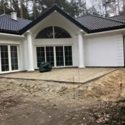 Przygotowanie podłoża pod taras z kostki przy białym domu z kolumnami, widoczne wytyczone granice i ubity piasek.