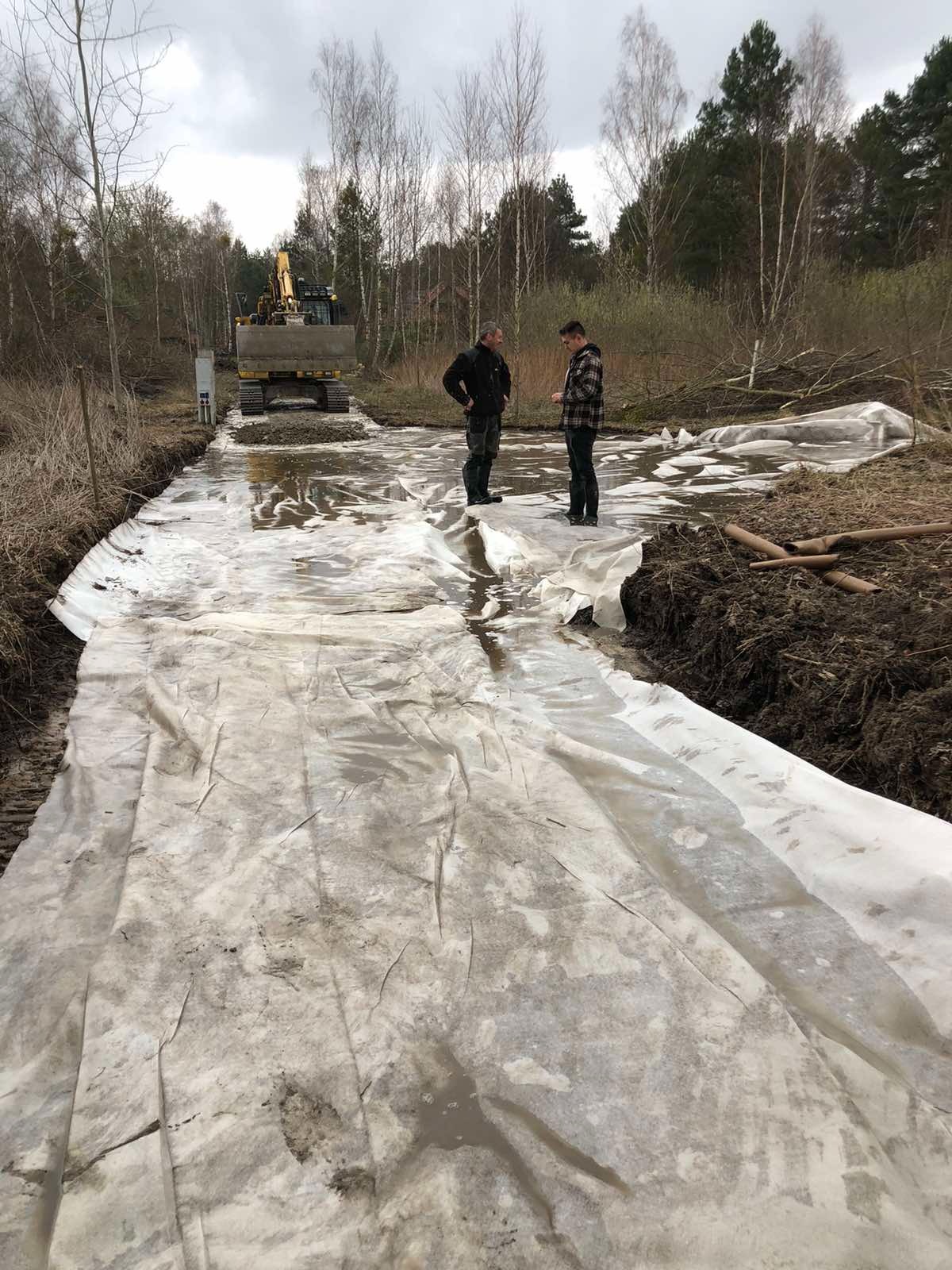Przygotowanie podłoża pod budowę drogi: rozłożona geowłóknina na błotnistym terenie, w tle koparka i dwóch mężczyzn omawiających plan działania.