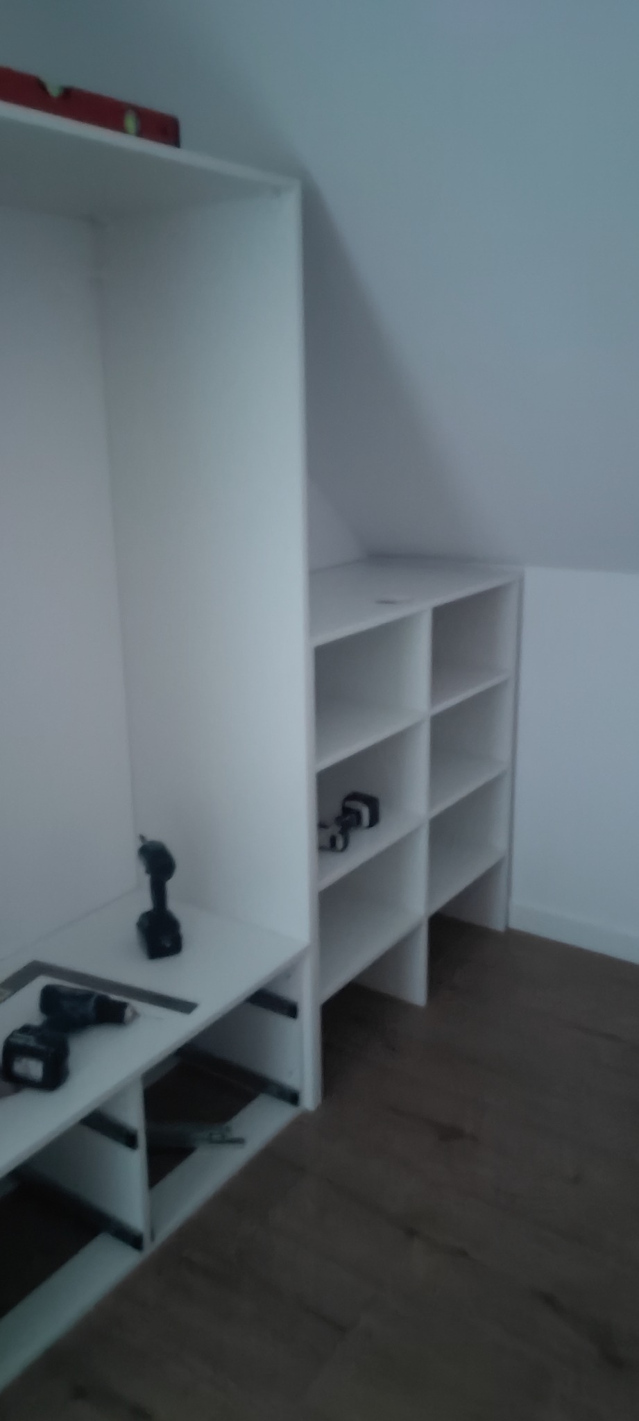 Biała szafa wnękowa z półkami pod skosem dachu, w trakcie montażu widoczne narzędzia: wkrętarki i poziomica.