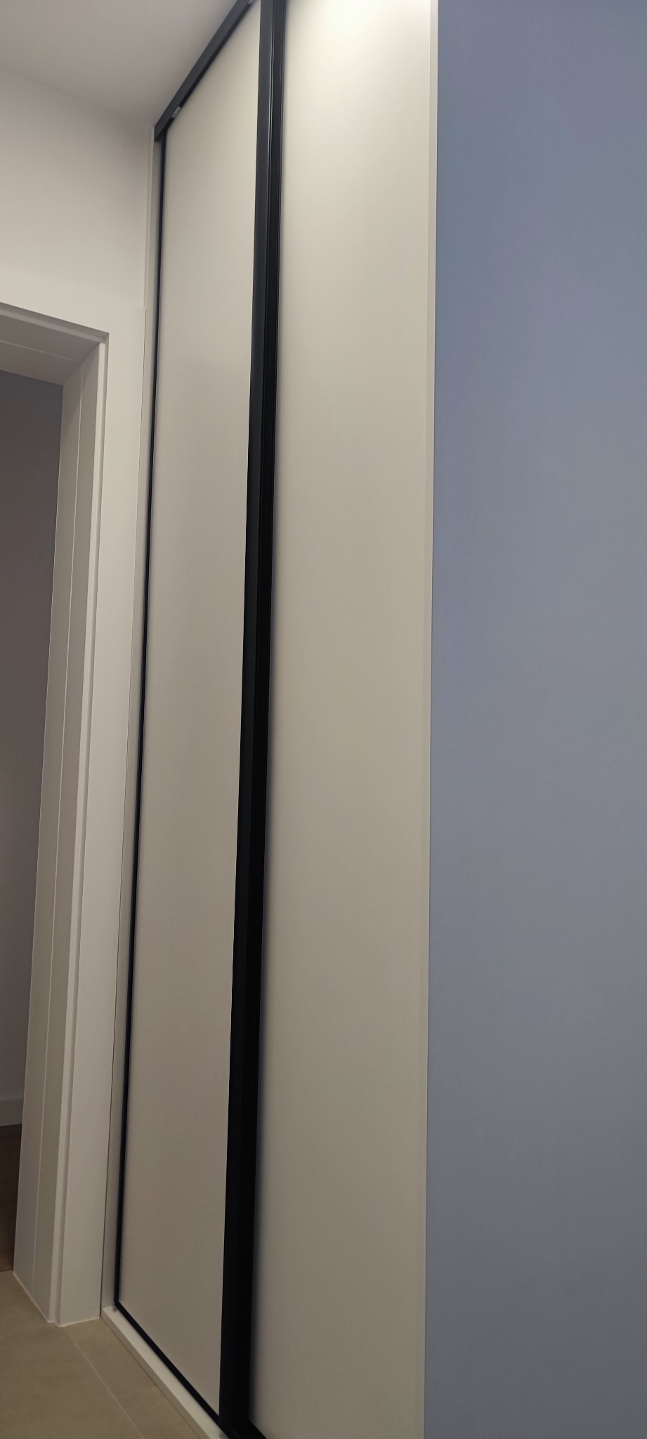 Zamknięta szafa wnękowa z przesuwnymi drzwiami w kolorze kremowym z czarnymi ramami, widoczna z boku na tle białej ściany i fragmentu ściany w kolorze gołębiej szarości.