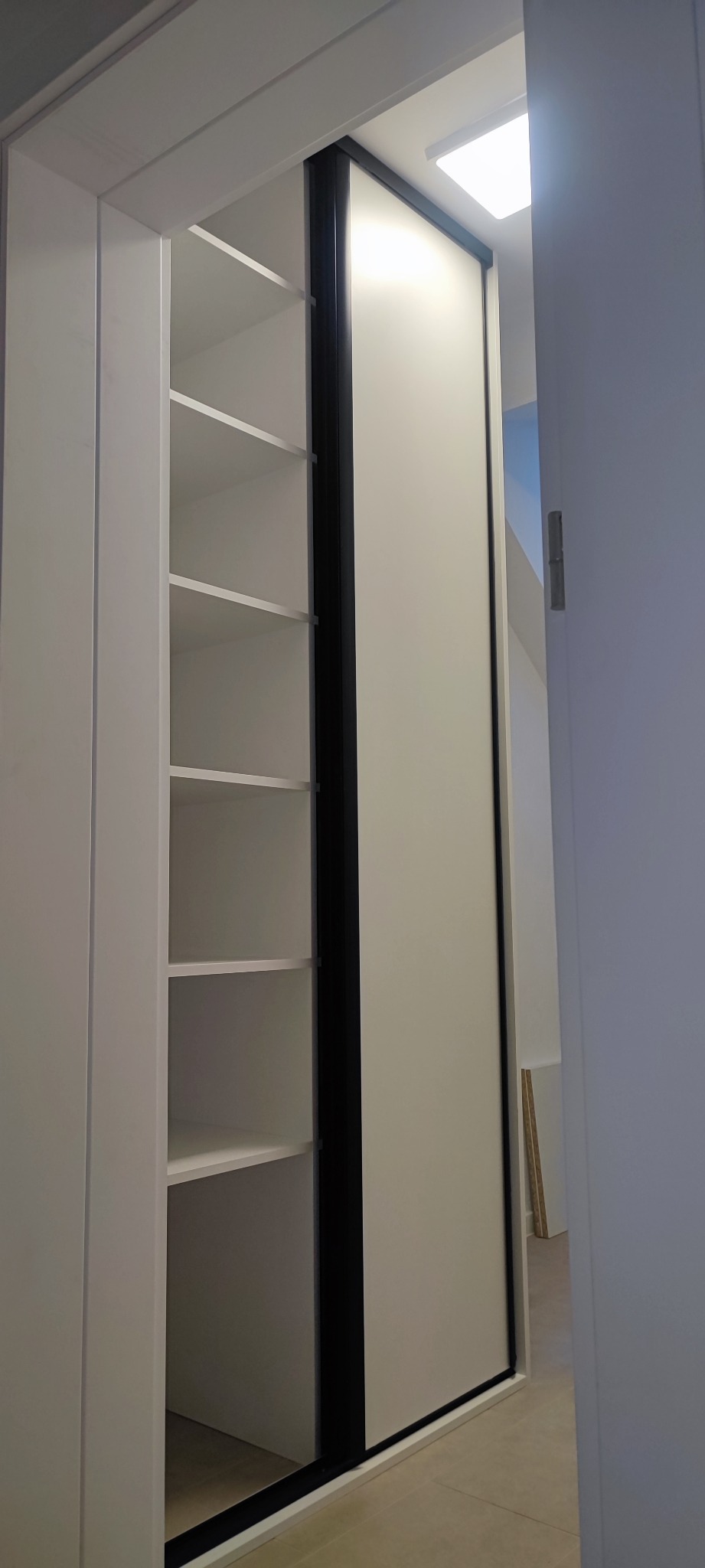 Biała szafa wnękowa z przesuwnymi drzwiami z czarnymi obramowaniami, widoczne półki wewnętrzne, minimalistyczny design.