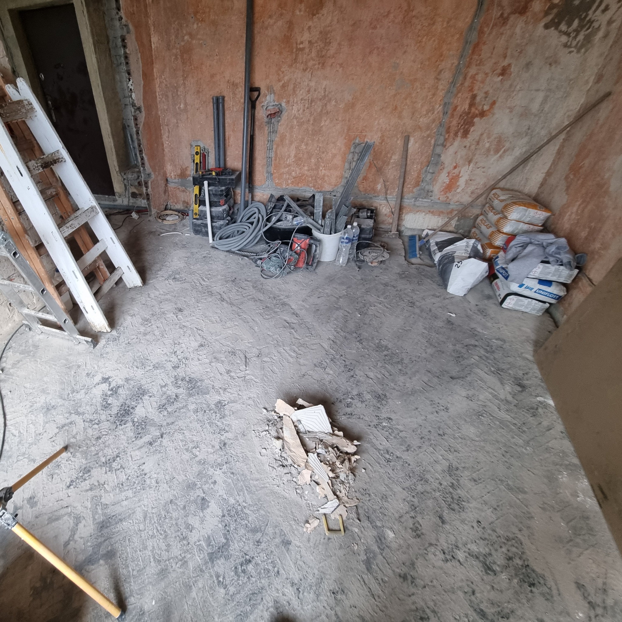 Wnętrze w trakcie remontu: narzędzia, drabina, worki z zaprawą i gruz na podłodze, ściany z widocznymi śladami prac murarskich.
