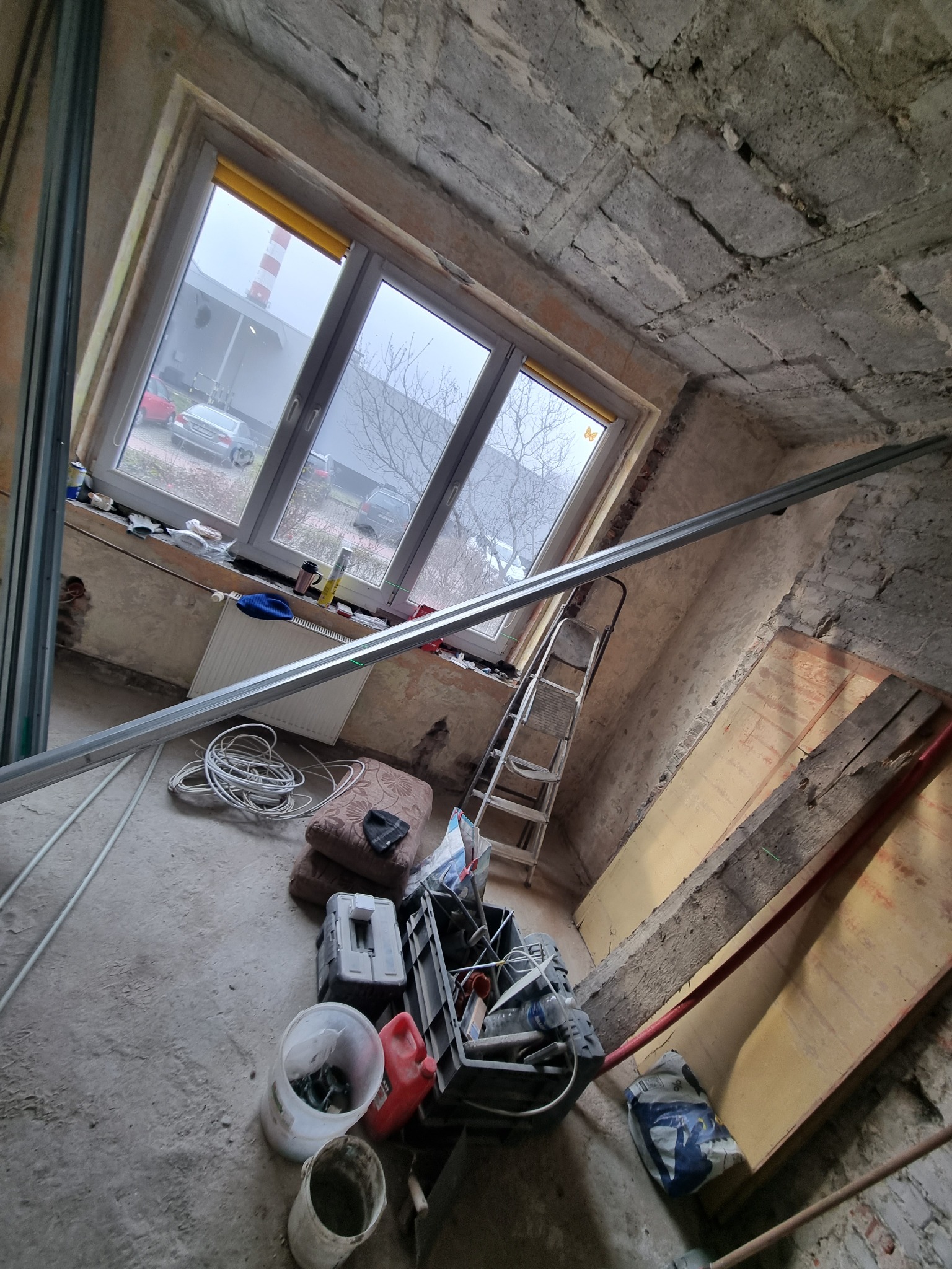 Wnętrze pokoju w trakcie remontu: surowy betonowy sufit, okno z żółtą roletą, drabina oparta o ścianę, profile metalowe, narzędzia i materiały budowlane na podłodze, widok na zewnątrz z kominem w tle.
