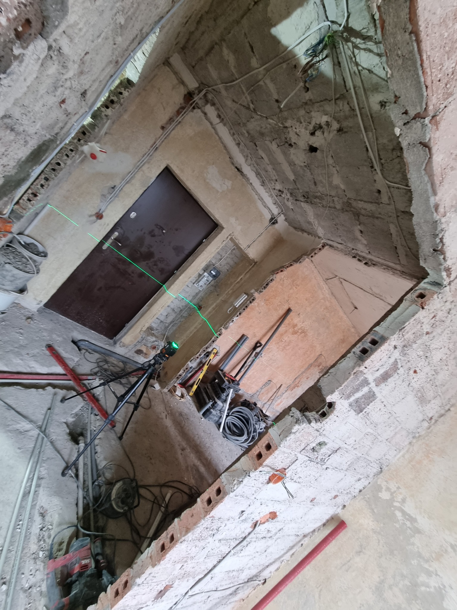 Wnętrze w trakcie prac remontowych, widoczne surowe ściany, instalacje, narzędzia i laser krzyżowy wyznaczający poziom, stan deweloperski.