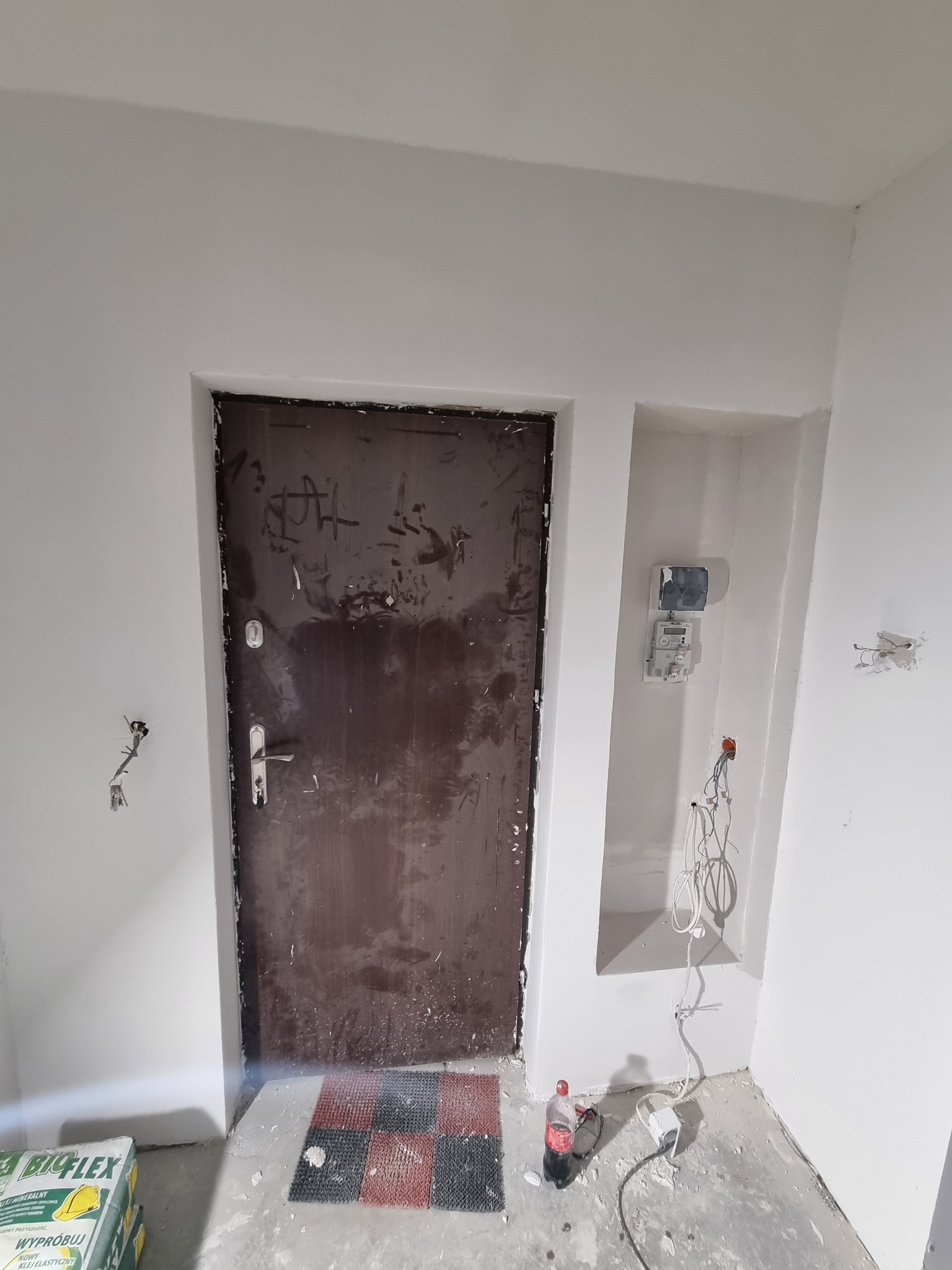 Wnętrze w trakcie prac remontowych: widoczne ślady po szpachlowaniu na ścianach, stara, zabrudzona brązowa futryna i drzwi wejściowe, wystające przewody elektryczne oraz licznik w niszy.