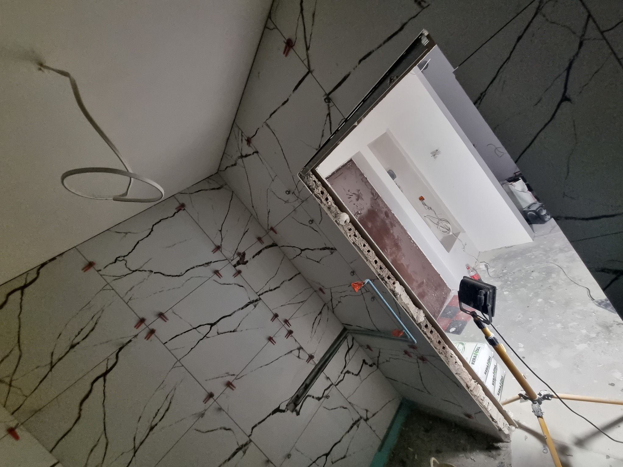 Montaż płytek imitujących marmur w łazience, widoczne krzyżyki dystansowe, lampa robocza i fragment instalacji hydraulicznej.