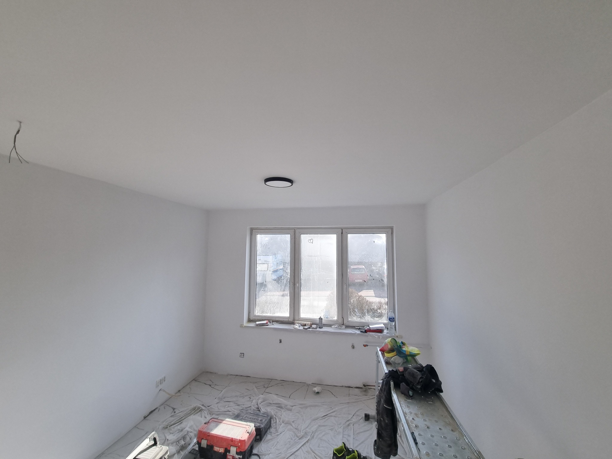 Wnętrze pokoju po malowaniu na biało, widoczne narzędzia malarskie, drabina i osłonięta folią podłoga, z wiszącymi kablami na suficie i okrągłym plafonem.