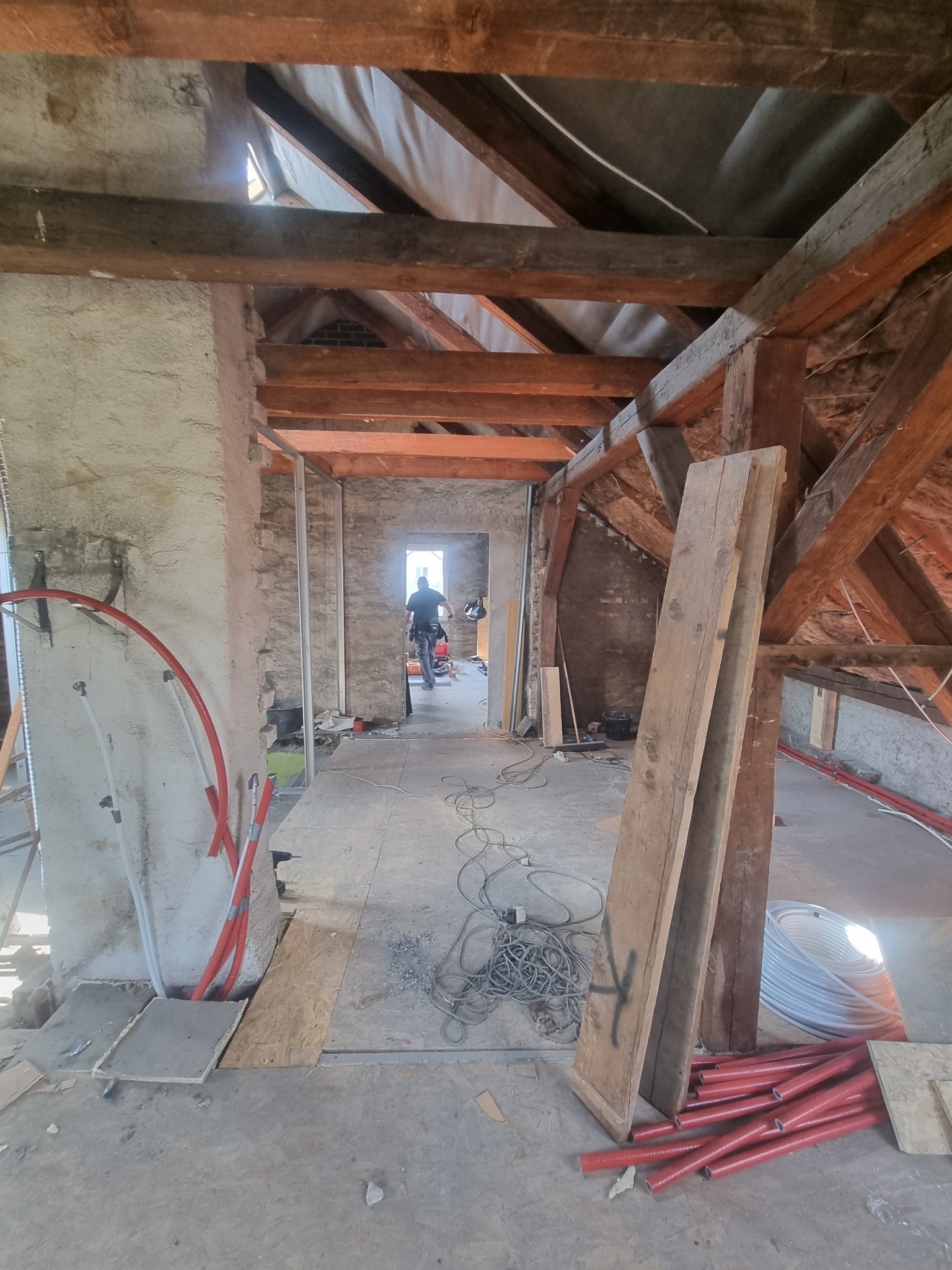 Poddasze w trakcie remontu: widoczne drewniane belki stropowe, fragmenty ścian z surowej cegły, instalacja grzewcza w czerwonych rurach oraz majster w oddali, pracujący w przejściu do kolejnego...