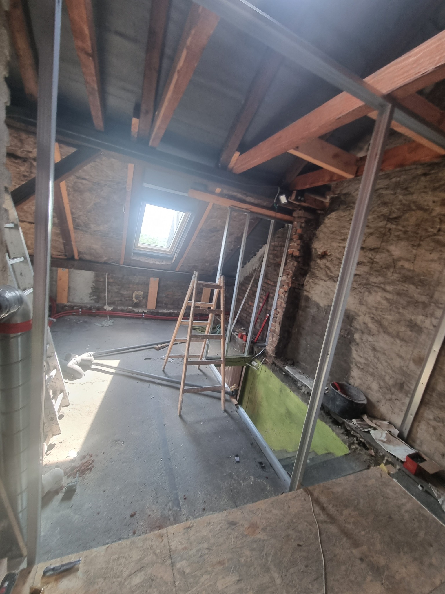 Poddasze w trakcie adaptacji: widoczne metalowe profile konstrukcyjne ścian działowych, drewniana drabina, okno dachowe wpadające światło, fragmenty starej ściany z cegły, instalacja elektryczna...
