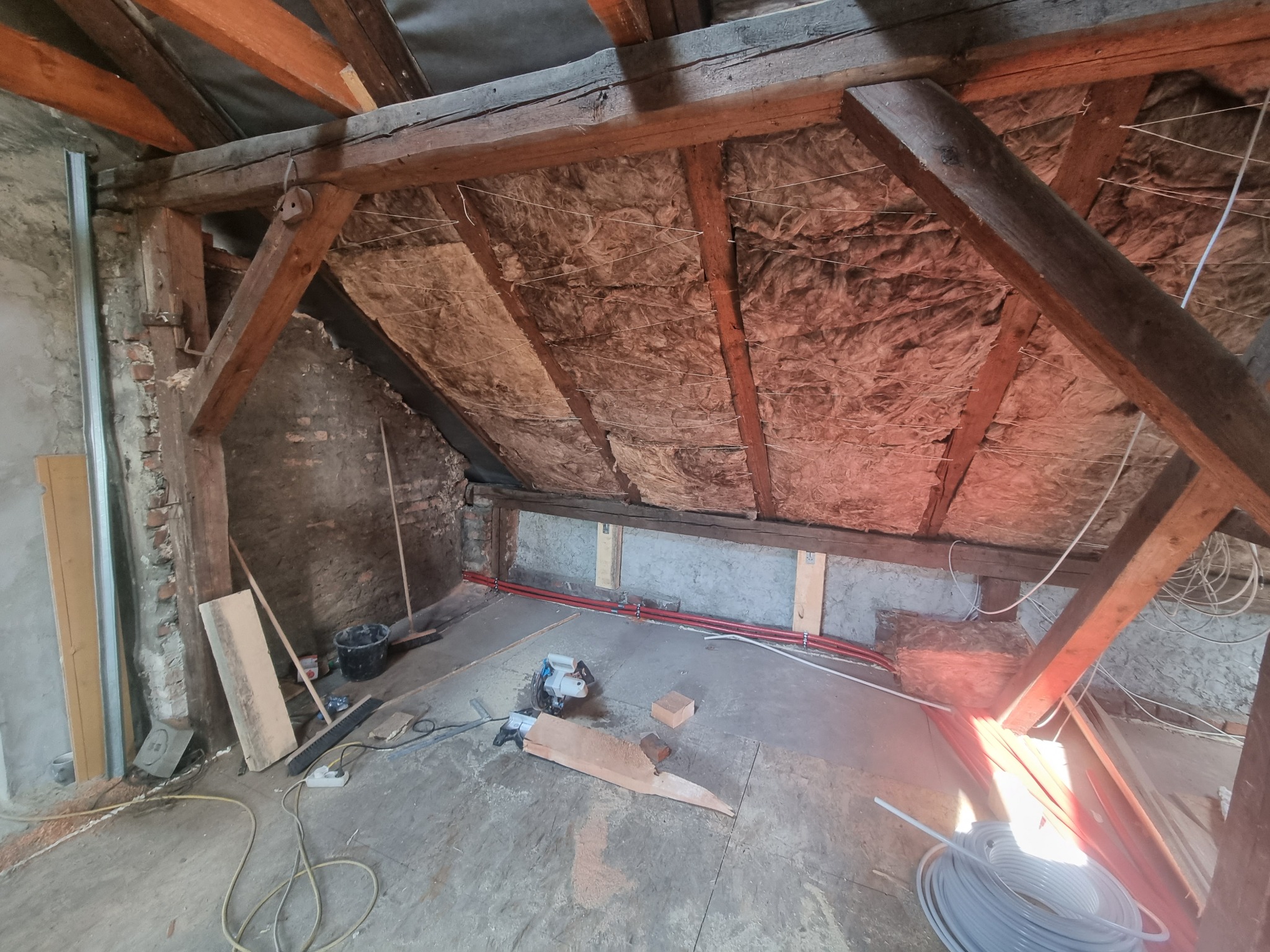 Poddasze w trakcie adaptacji: widoczne ocieplenie dachu wełną mineralną, drewniana konstrukcja więźby dachowej, fragmenty instalacji grzewczej, narzędzia i materiały budowlane na podłodze, surowe...