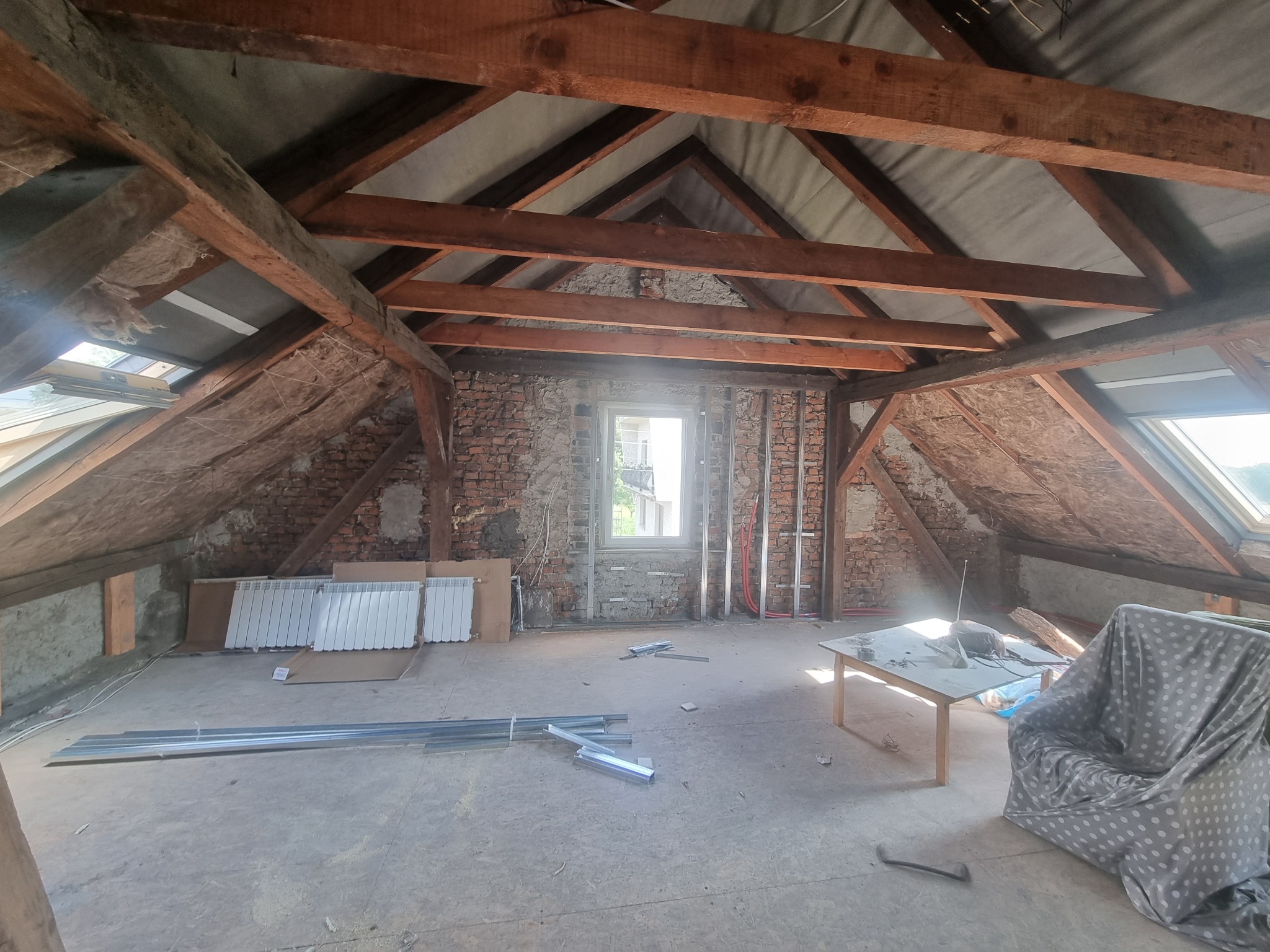 Poddasze w trakcie adaptacji: widoczne drewniane belki stropowe, surowa cegła, okno dachowe, grzejniki, profile metalowe, częściowo obita ściana i meble przykryte folią.