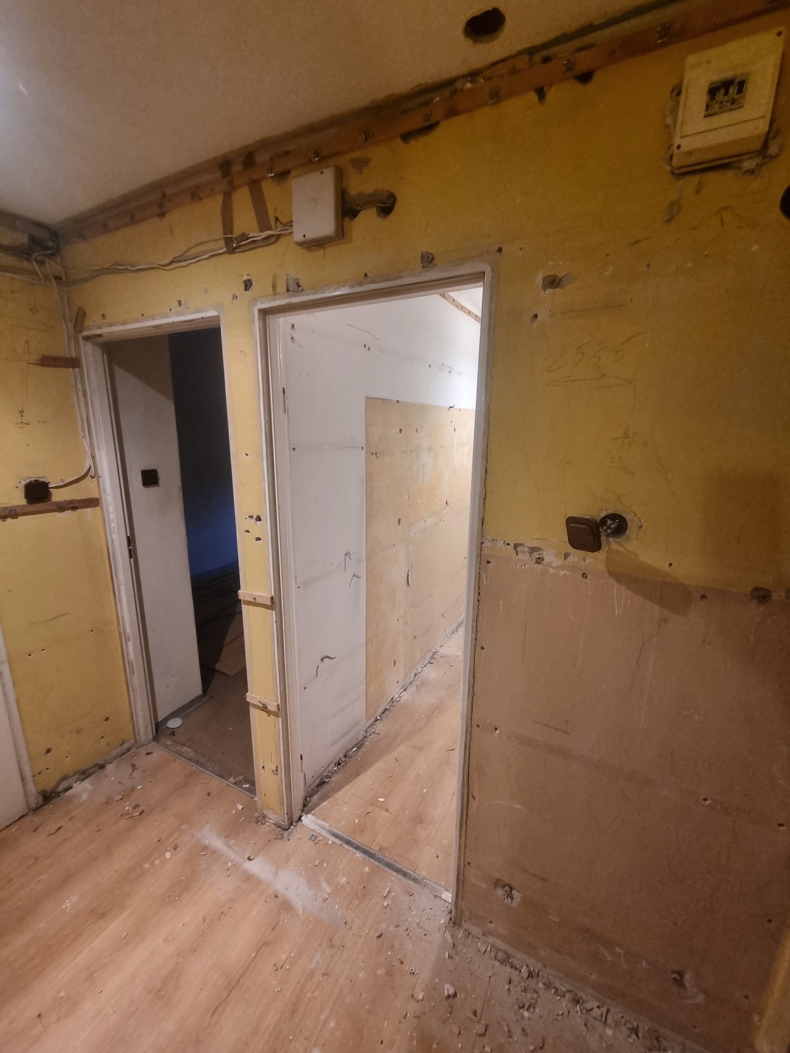 Wnętrze mieszkania w trakcie remontu: odsłonięte przewody elektryczne, fragmenty starej tapety na żółtej ścianie, przygotowane otwory pod gładzie, widoczne dwie futryny drzwiowe.