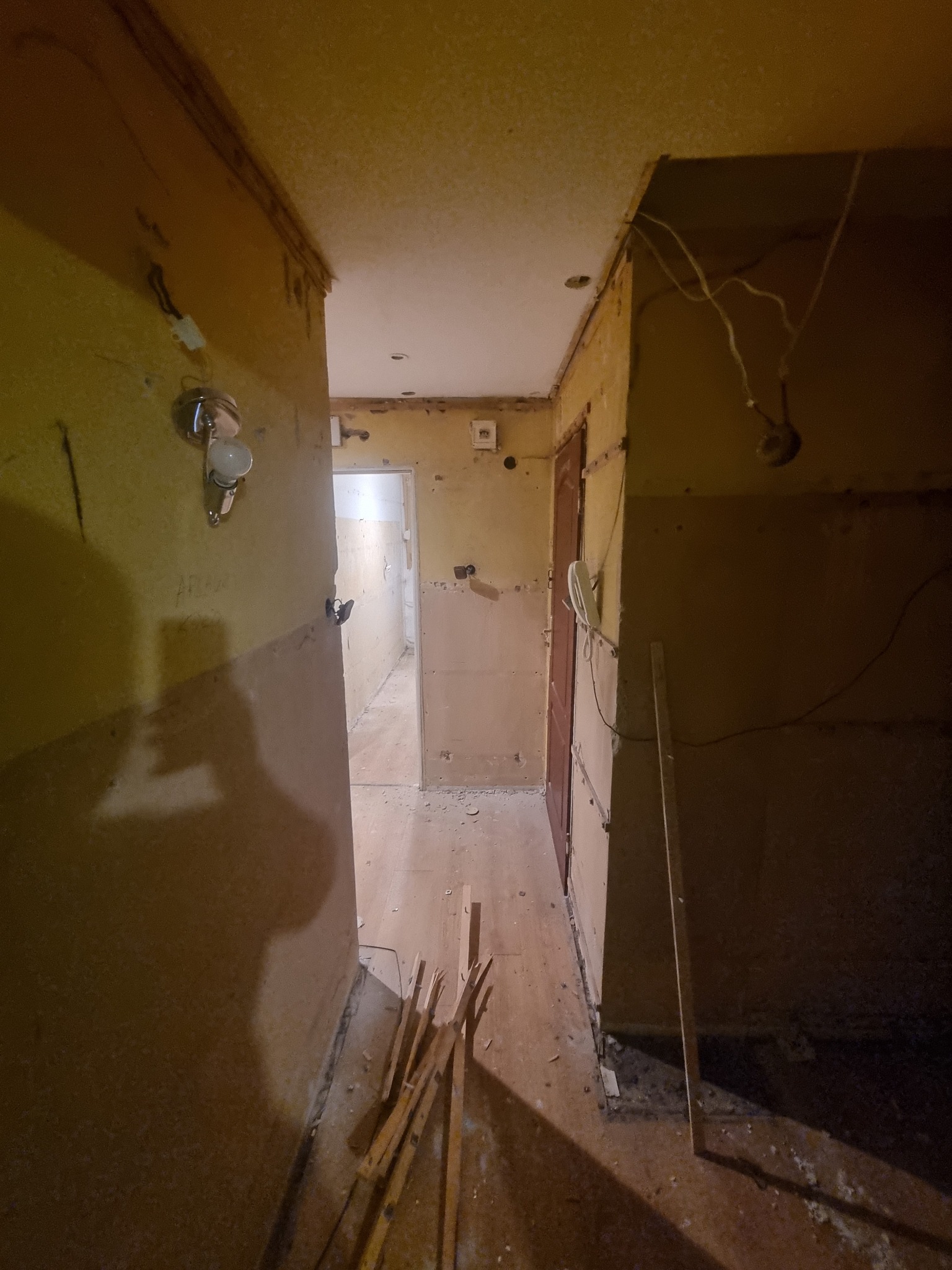 Korytarz w trakcie remontu: zerwane tapety, widoczne kable, stare drzwi, fragmenty listew przypodłogowych na podłodze.