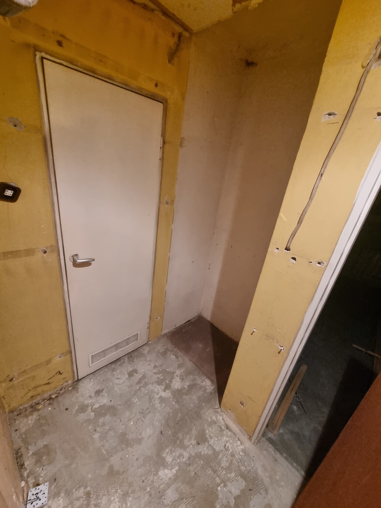 Wnętrze w trakcie remontu: ściany przygotowane do malowania, widoczne ślady po instalacjach elektrycznych, surowy beton na podłodze, białe drzwi z kratką wentylacyjną w dolnej części.