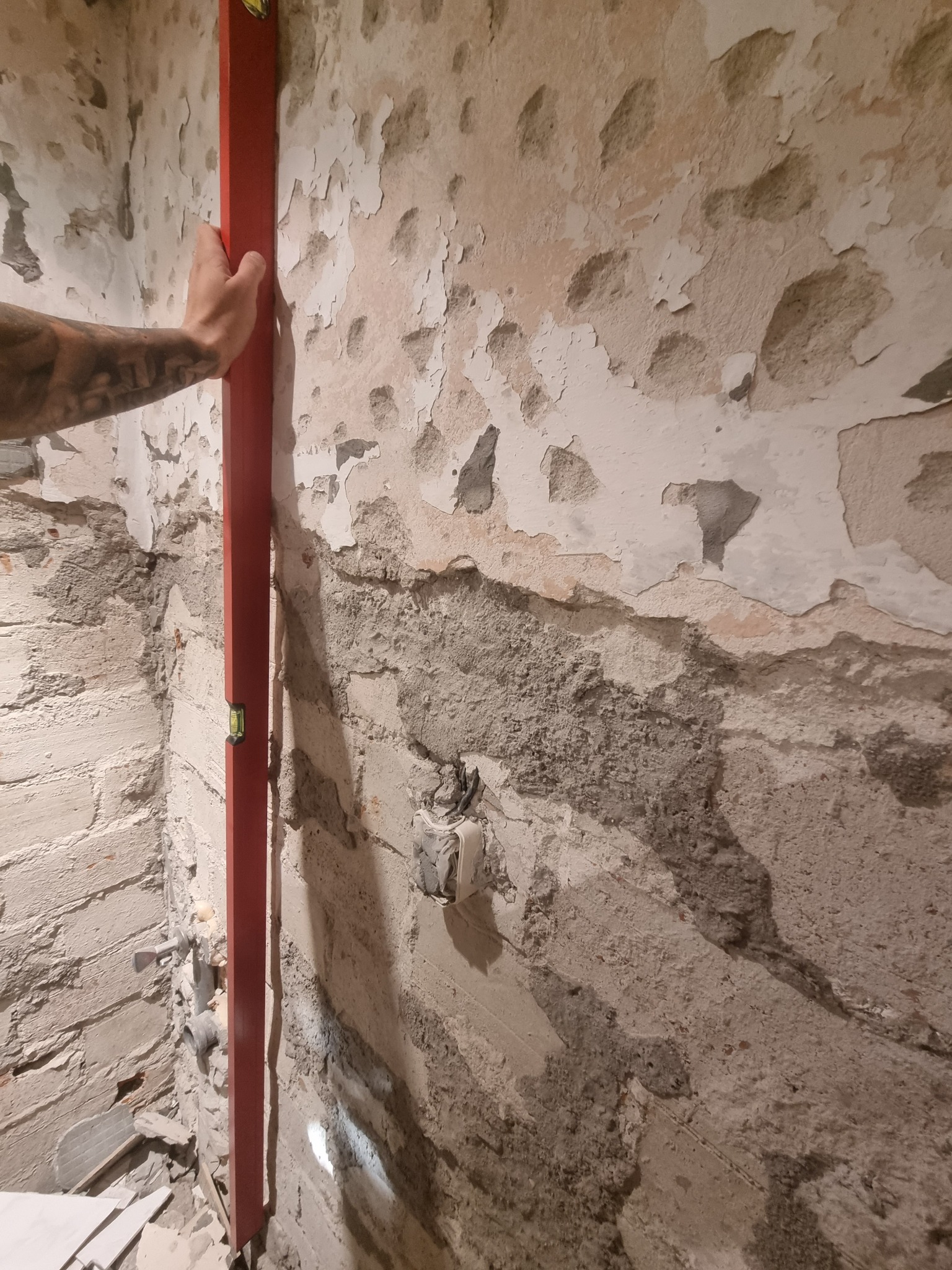 Ręka z tatuażem przytrzymuje czerwoną poziomicę na nierównej ścianie z odpadającą farbą, widoczne stare warstwy tynku i instalacja elektryczna.