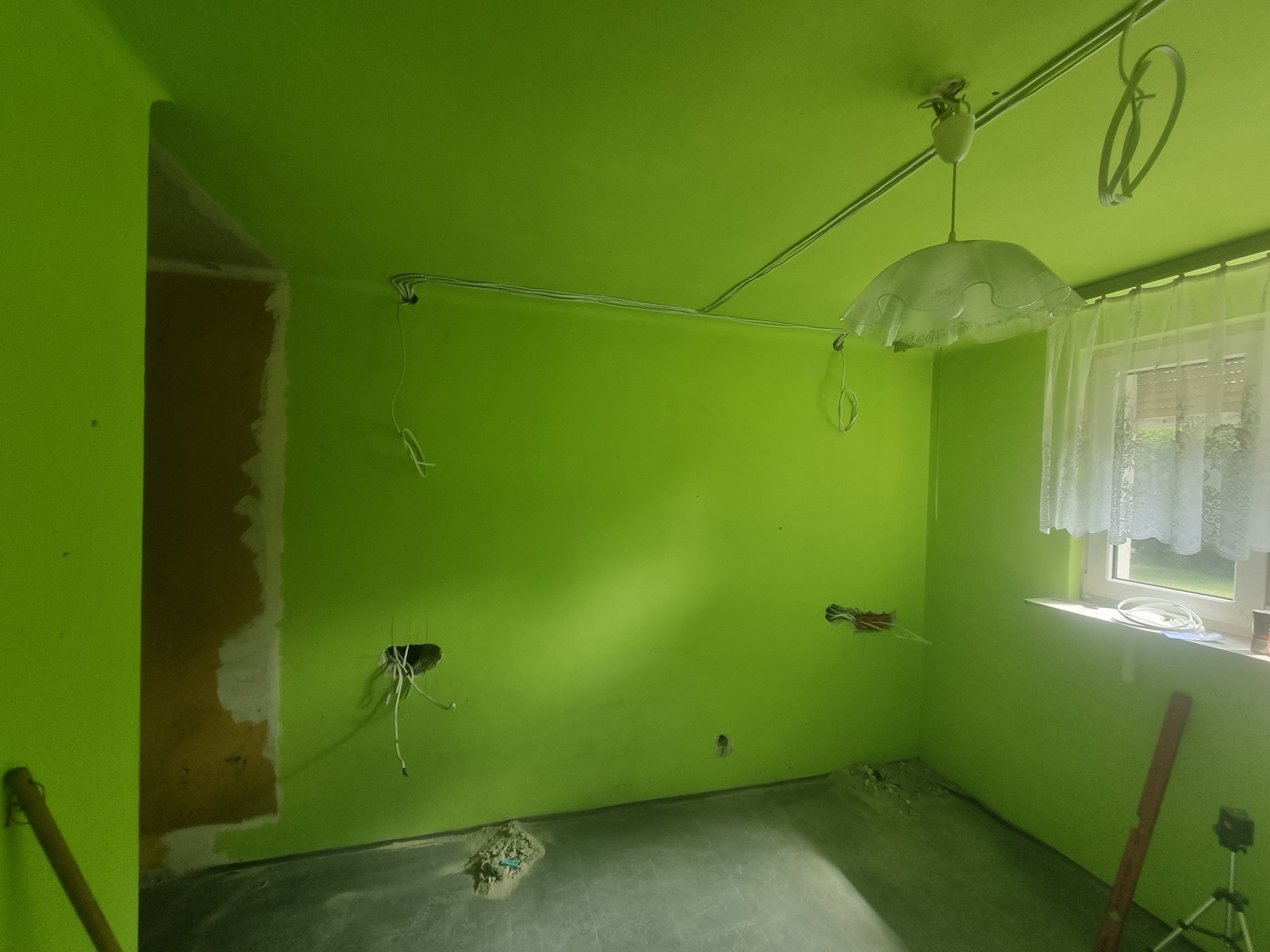 Pokój pomalowany na jaskrawy zielony kolor w trakcie remontu, widoczne wystające kable ze ścian i sufitu, przygotowane otwory pod gniazdka, stara lampa sufitowa, okno z firanką, resztki gruzu...