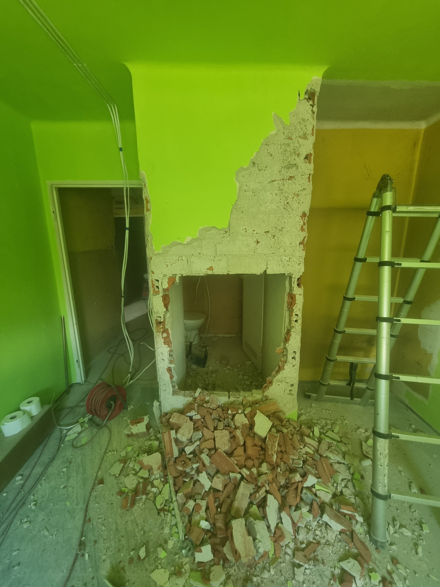 Wyburzanie ściany w łazience z widoczną toaletą w tle, gruz ceglany na podłodze, drabina oparta o ścianę, ściany pomalowane na jaskrawy zielony i żółty kolor.