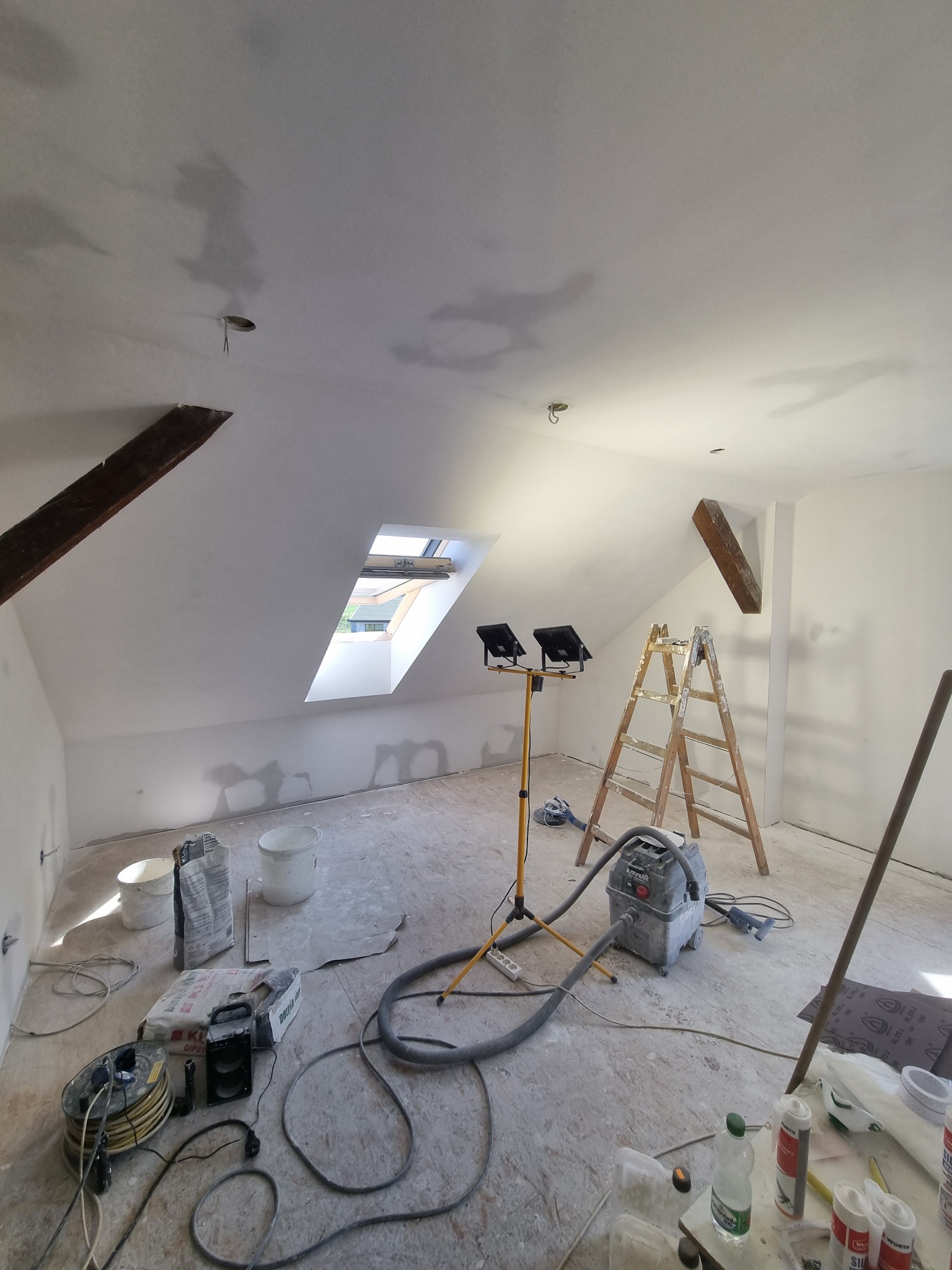 Poddasze w trakcie prac wykończeniowych: widoczne drewniane belki, okno dachowe, ściany i sufit po szpachlowaniu, drabina, lampy budowlane oraz narzędzia i materiały budowlane na podłodze.