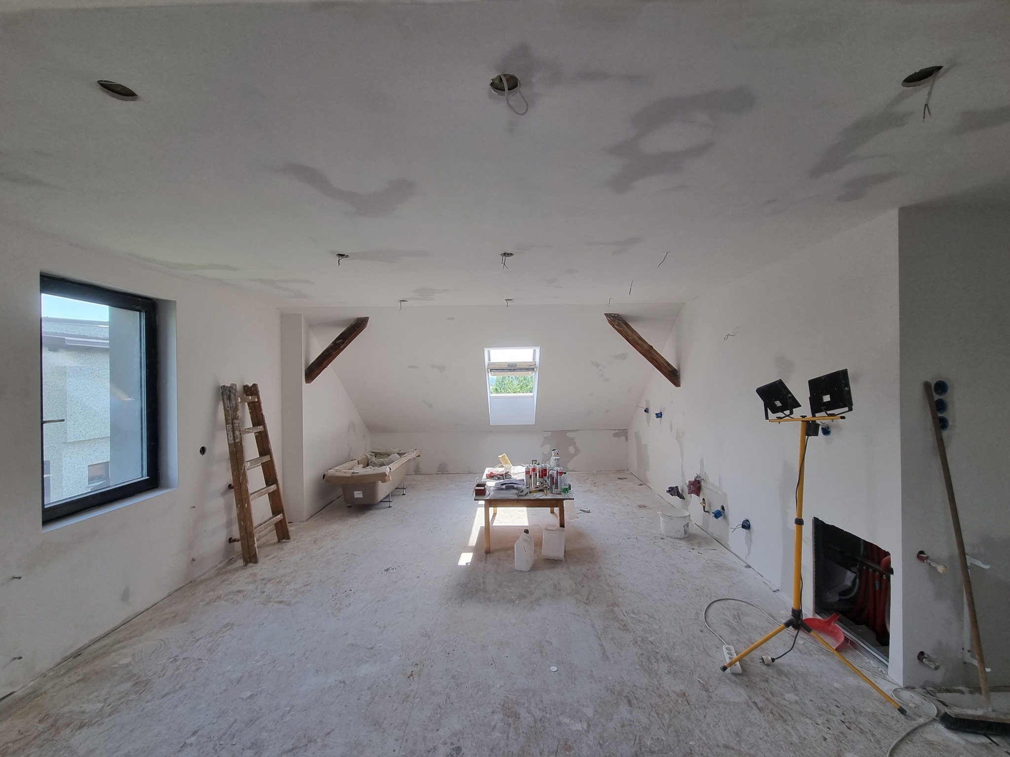 Poddasze w trakcie remontu: świeżo wygładzone ściany, sufit z otworami na oświetlenie, drabina oparta o ścianę przy oknie, stół z narzędziami i materiałami malarskimi pod oknem dachowym, lampa...