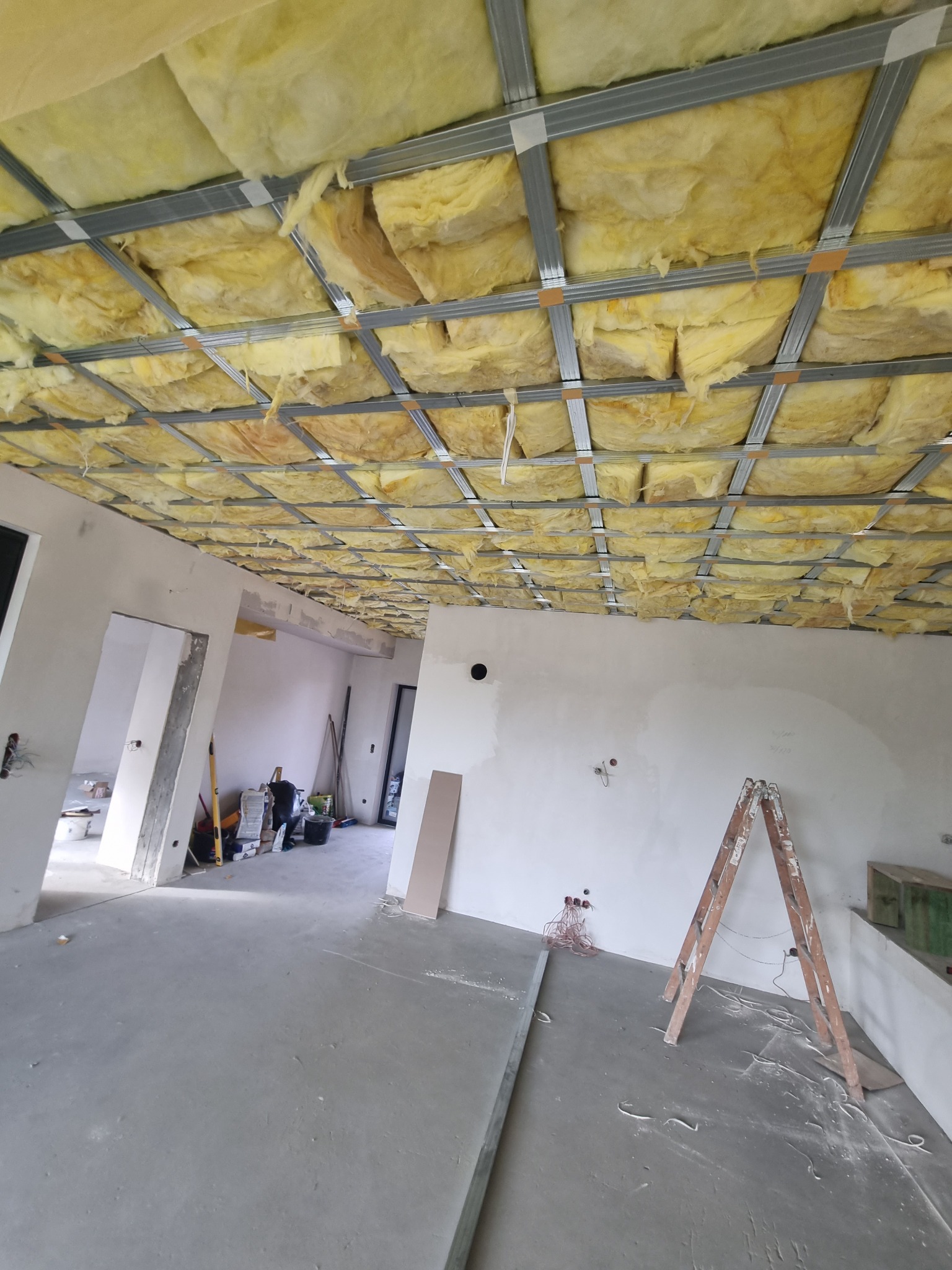 Montaż izolacji z wełny mineralnej w metalowej konstrukcji sufitu podwieszanego w surowym wnętrzu, widoczna drabina oparta o ścianę.